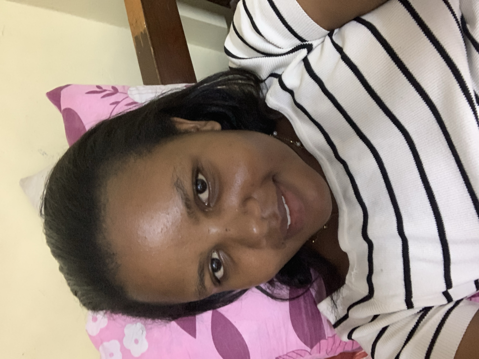 A person is lying on a pink pillow with a floral pattern. The person is wearing a white shirt with black stripes. The person has dark hair and dark eyes. The location is not evident in the image.