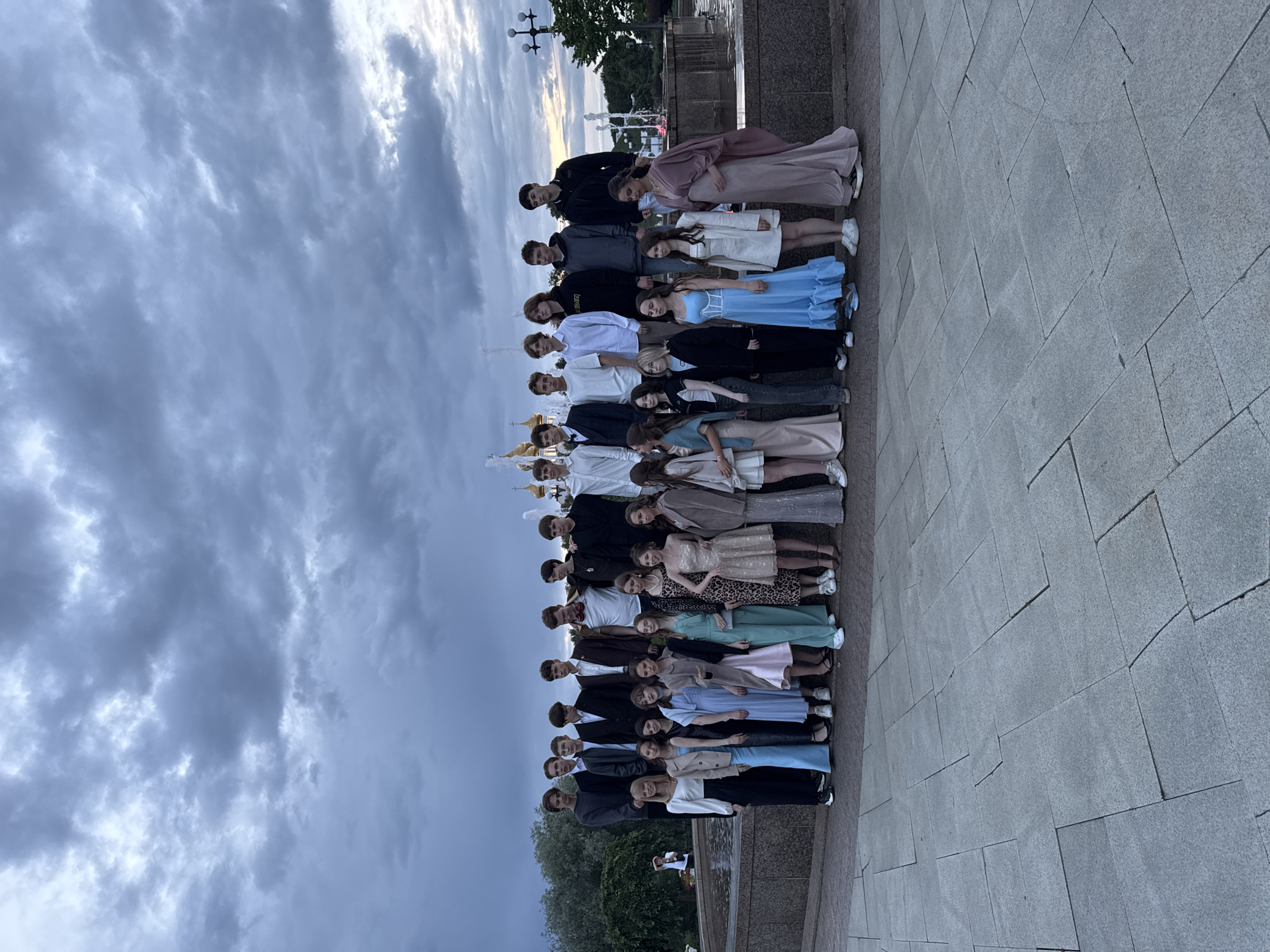 This media file captures a group of young people, likely students, posing for a photograph in what appears to be an outdoor public space. The group is arranged in a staggered line, with individuals standing close together, suggesting a formal or celebratory occasion. The subjects are dressed in a mix of formal attire, with many of the young women wearing long dresses in various colors, including pale pink, blue, and light brown, while the young men are in suits and casual wear.
The setting features a paved walkway or plaza made of large, rectangular stone tiles. In the background, a fountain can be seen with water spraying, and beyond that, trees and what looks like the facade of a building, possibly with golden domes, hinting at a park or urban landmark. The sky is cloudy and overcast, suggesting it might be evening or that the weather is changing. The light appears diffused, contributing to a slightly muted color palette.
The arrangement of the people and their attire implies a gathering like a prom, graduation ceremony, or a similar special event. The overall mood seems composed, with most individuals looking towards the camera. There is no visible text in the image. The image is oriented vertically, with the sky at the top and the ground at the bottom, though the subjects are also oriented vertically within the frame.