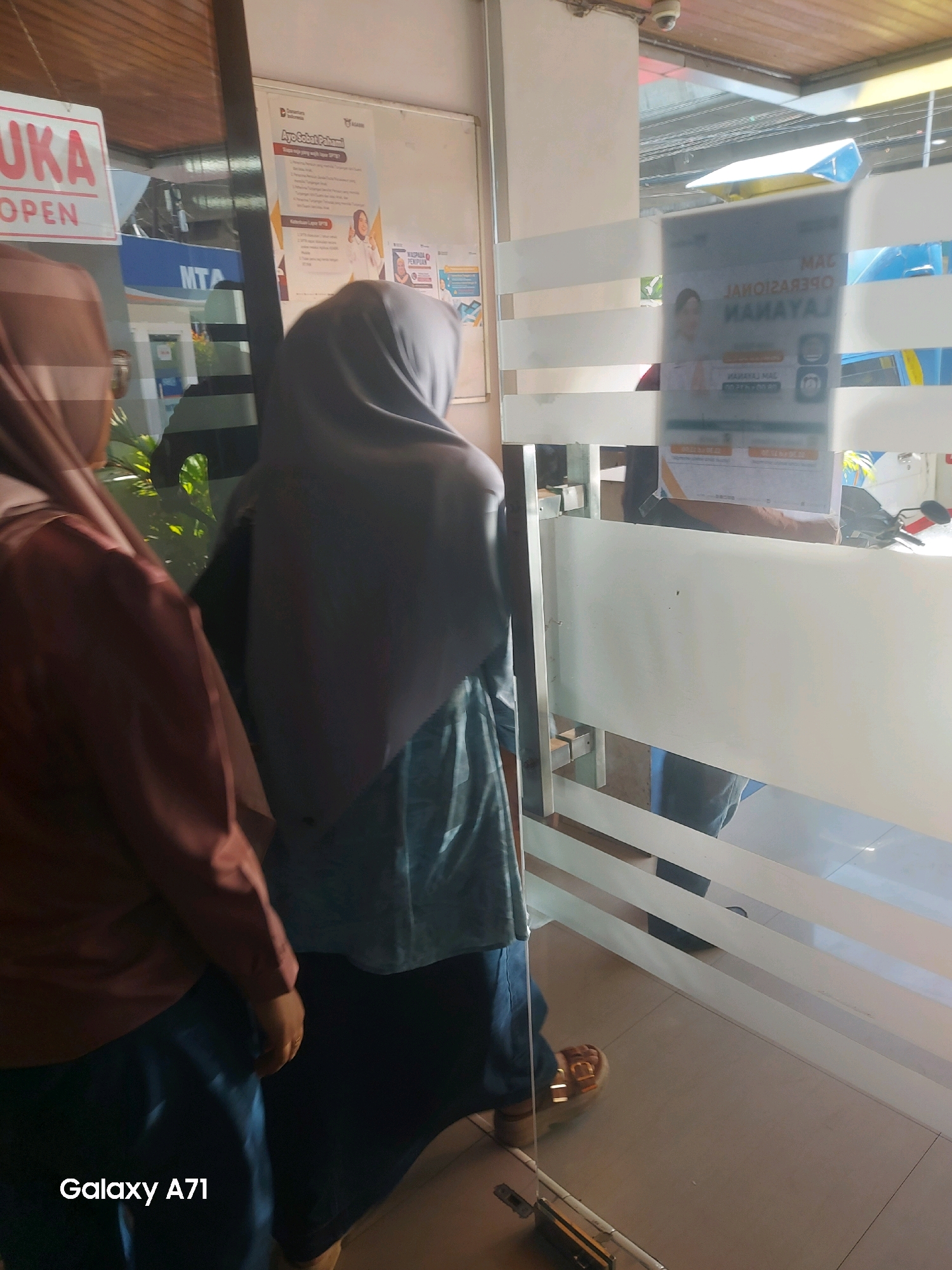 Two women are standing inside a building, near a glass door. The woman on the left wears a brown headscarf and a brown shirt, and the woman on the right wears a grey headscarf and a blue and grey patterned dress. They are both facing away from the camera, looking into the building. A sign with red lettering that says "BUKA" and "OPEN" is visible through the glass door on the left. On the right side of the image, a poster with Indonesian text is attached to the glass door. The poster appears to be a schedule of operating hours, with text such as "JAM OPERASIONAL" and "LAYANAN". The floor is tiled. In the background, there are glimpses of vehicles and buildings, suggesting an urban environment. The lower-left corner of the image shows the text "Galaxy A71".