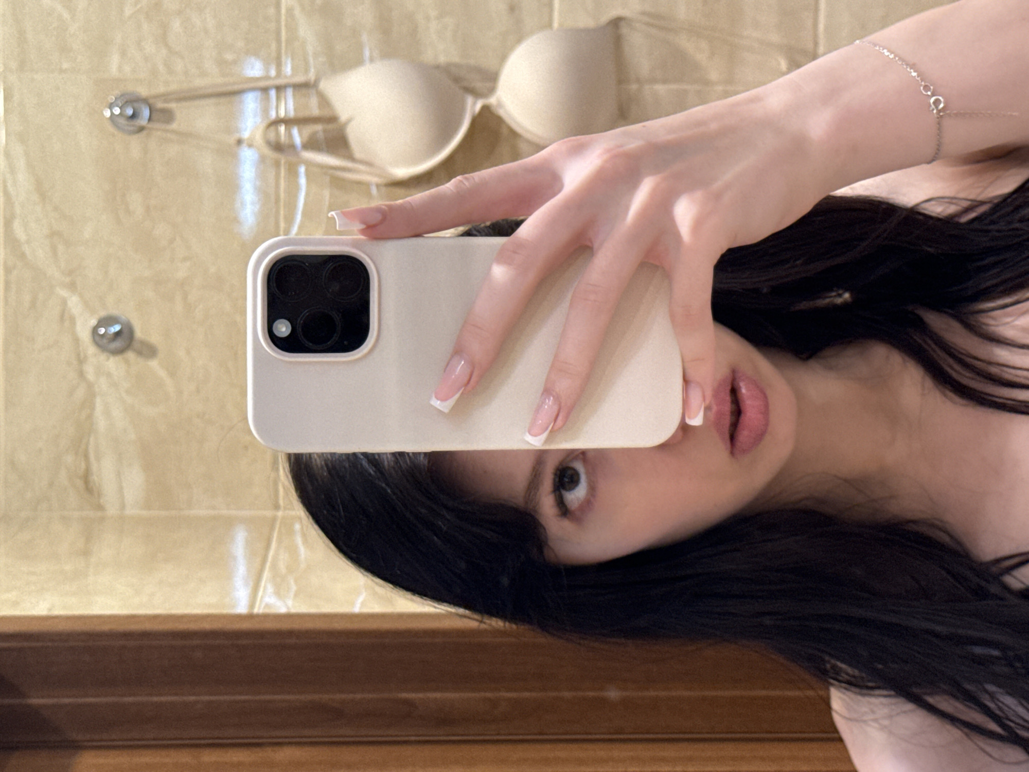 A young woman is taking a selfie in a bathroom. She is holding a light-colored smartphone in her right hand, obscuring the center of her face. Her dark hair frames the visible portions of her face, which include her left eye and lips. The background consists of beige tiled walls with two chrome hooks on the right side. A nude bra is hanging from one of the hooks. A wooden-framed mirror is visible to the left of the woman. She is wearing a delicate silver bracelet on her left wrist.
