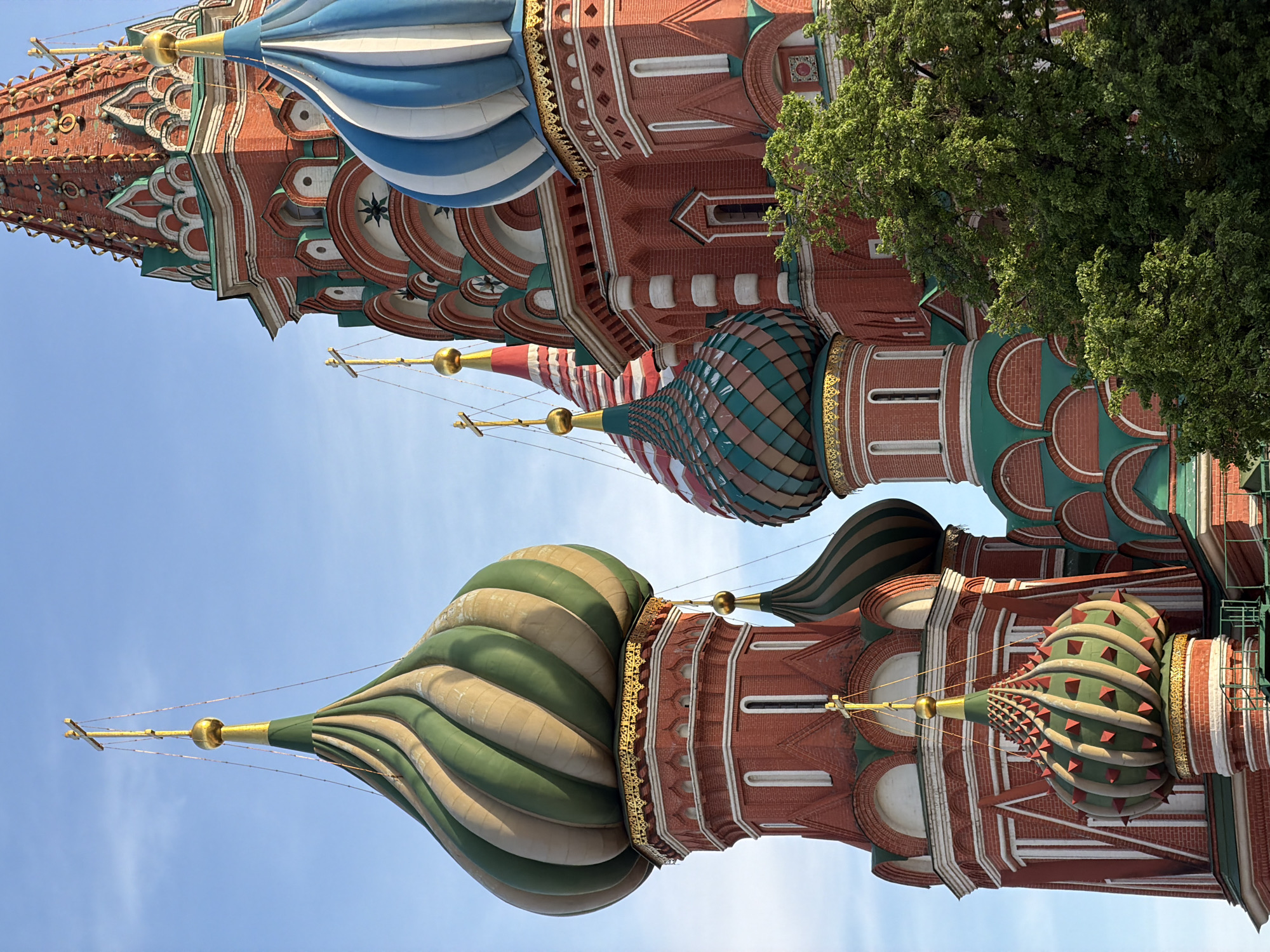 The image depicts a partial view of St. Basil's Cathedral, located in Moscow, Russia. The perspective is from below, angled upwards towards the sky, capturing several of the cathedral's iconic onion domes and architectural details. The domes feature varied colors and patterns, including blue and white stripes, green and tan swirls, and green with red triangular accents. The walls of the cathedral are predominantly red brick, adorned with decorative white and green elements. A green tree is visible to the right. The sky is a clear, light blue.