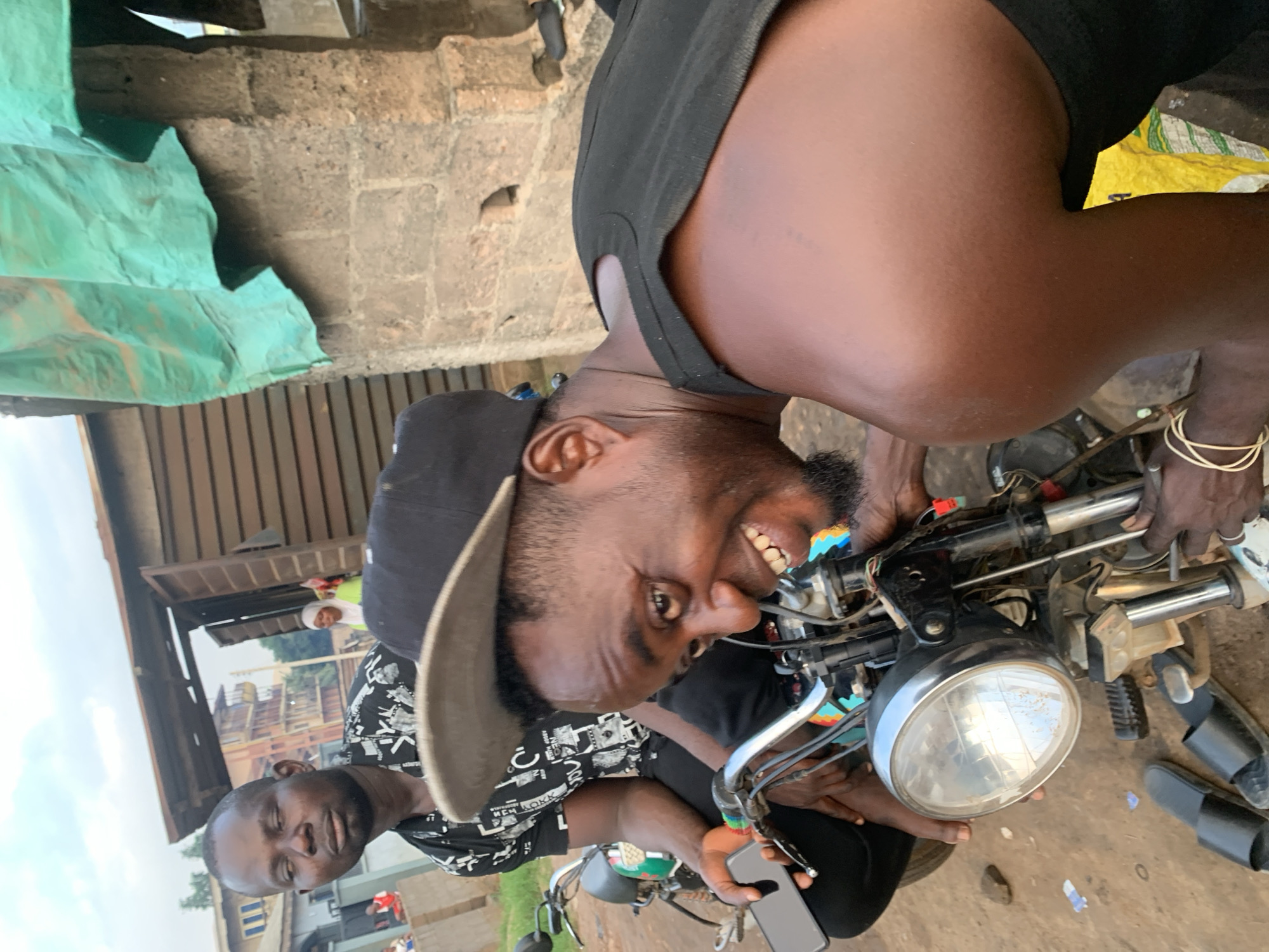 In Shagamu, Nigeria, two men are seated on a motorcycle. The man in the foreground, wearing a black tank top and a dark baseball cap, is leaning forward and smiling broadly at the camera. He has a beard and is holding onto the motorcycle's handlebars, one hand near the front brake lever. A second man, wearing a black and white patterned t-shirt, is seated behind him, looking away from the camera with a neutral expression. He is holding a black smartphone in his left hand. The motorcycle is parked on a dirt ground, with some debris and a pair of black sandals visible near its wheels. In the background, a wooden structure with an open doorway is visible, and beyond that, multi-story buildings and trees suggest an urban environment. A large piece of green tarp hangs in the upper right portion of the image.