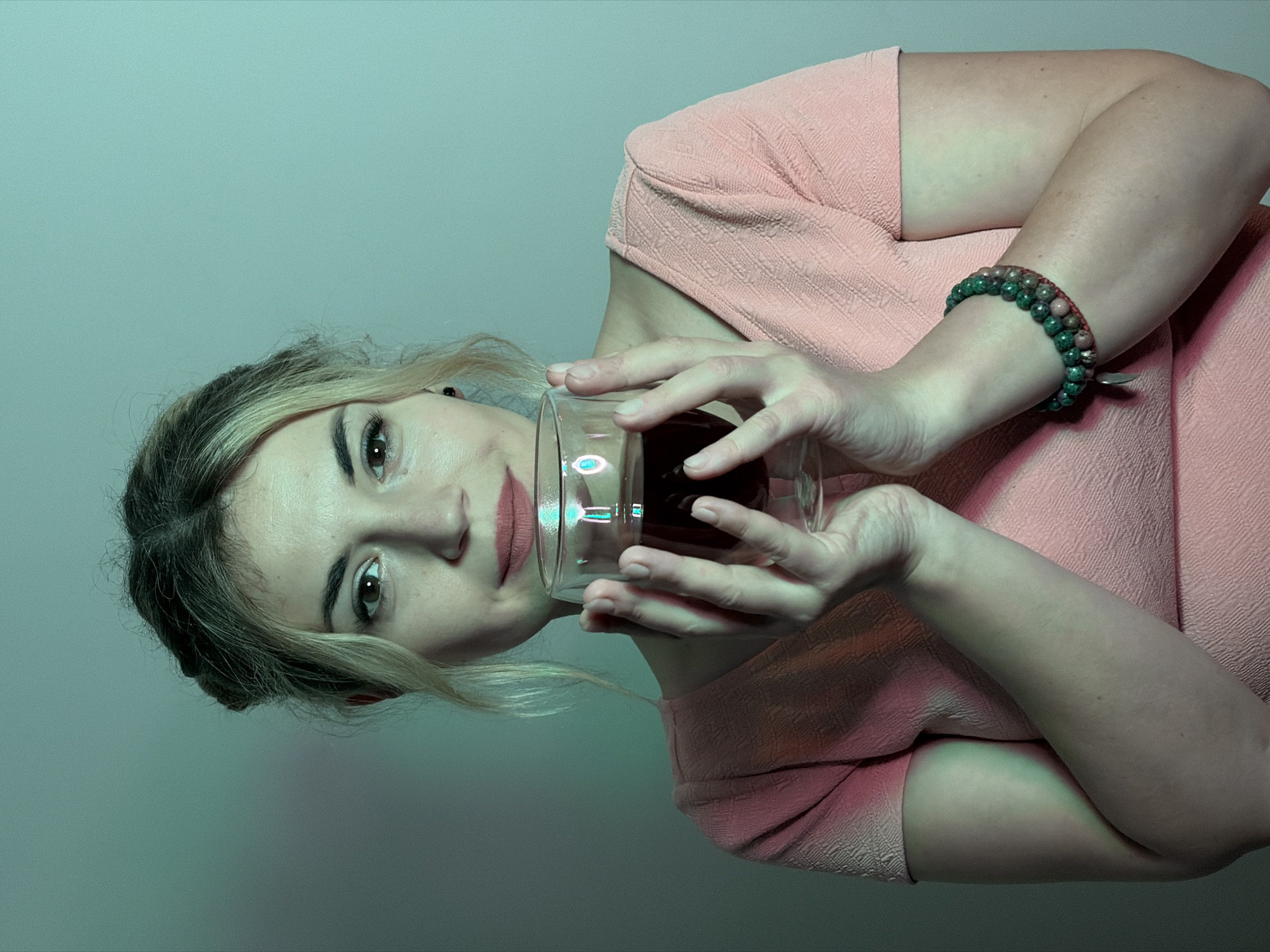 A woman is depicted looking directly at the camera, holding a clear glass filled with a dark red liquid, likely wine, in both hands. She has light-colored hair with darker roots, framing her face, and wears light pink lipstick. Her expression is calm and direct, with a hint of a subtle smile. She is dressed in a light pink, short-sleeved garment, possibly a dress or top, which features a prominent textured, geometric pattern. On her right wrist, she wears a multi-stranded beaded bracelet with beads in shades of green and reddish-pink. The background is a plain, solid light teal or greenish-blue color, indicating an indoor setting. The scene is illuminated by artificial lighting, creating a cool, ambient greenish-blue color cast across the entire image. Bright reflections are visible on the surface of the glass she holds. The overall aesthetic is stylized due to the distinctive lighting. This scene takes place in Budva, Montenegro.