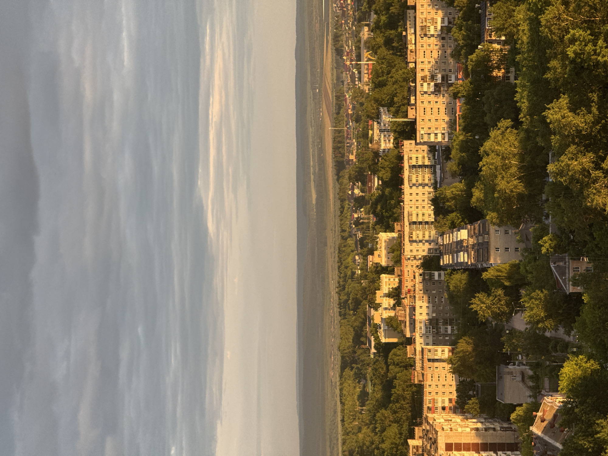 The image shows an aerial view of Perm, Russia, looking out from a high vantage point. The sky is overcast with a mix of gray and bright clouds. A wide expanse of densely wooded terrain stretches between the cityscape and the sky. The urban area comprises a dense collection of multi-story apartment buildings interspersed with trees. The buildings are light in color, likely concrete, with multiple windows. The sun appears to be setting, casting a warm glow on the buildings and trees. There are no visible people or actions. The image suggests a residential area in Perm near a wooded periphery.