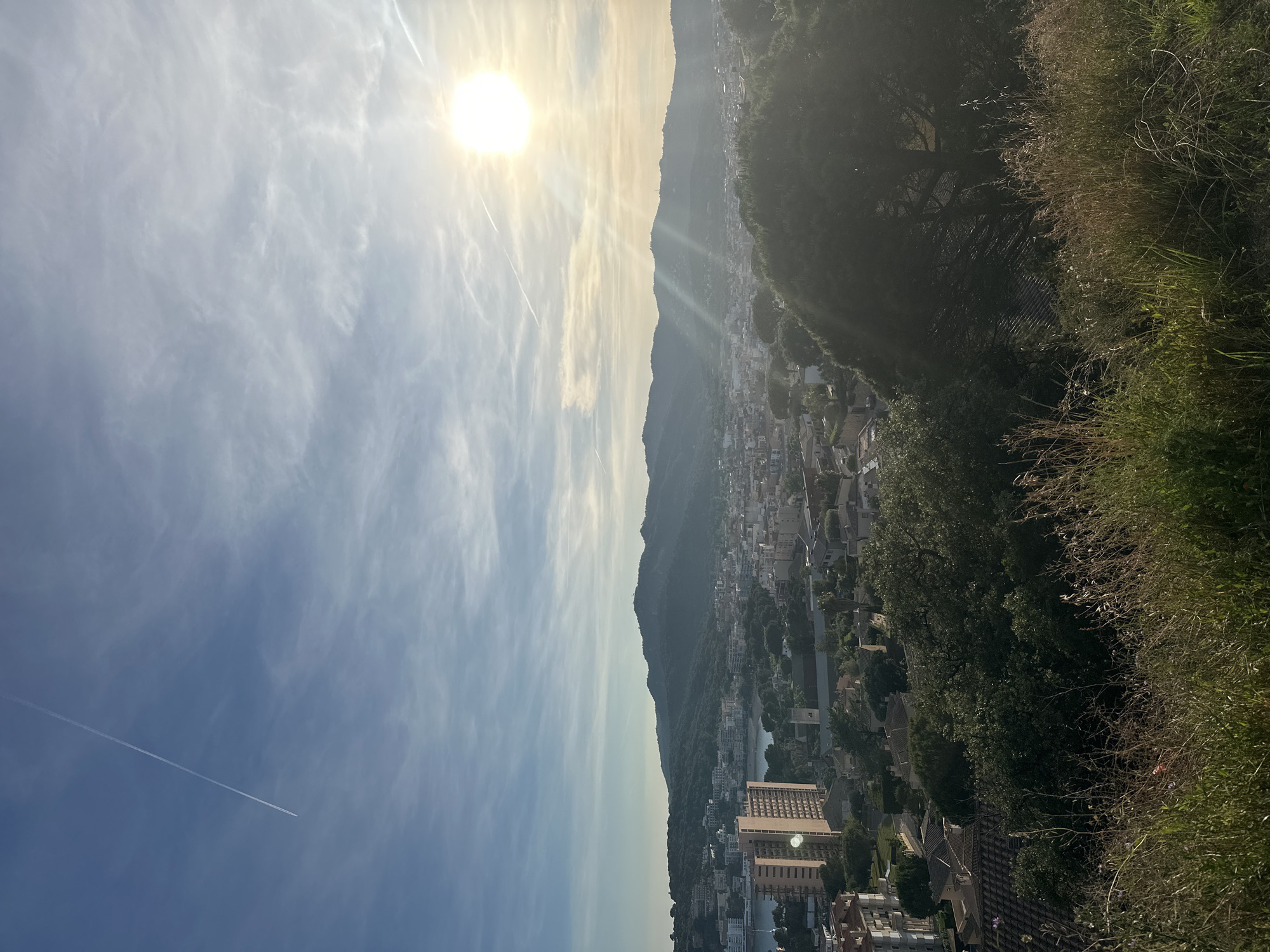 The image captures a panoramic view of Sant Feliu de Guíxols, Spain, from an elevated vantage point. The scene is dominated by a landscape with buildings, trees, and hills. The sky is clear with a few clouds and contrails, and the sun is shining brightly, casting a warm glow over the landscape. The vegetation is abundant, adding a lush green texture to the composition.