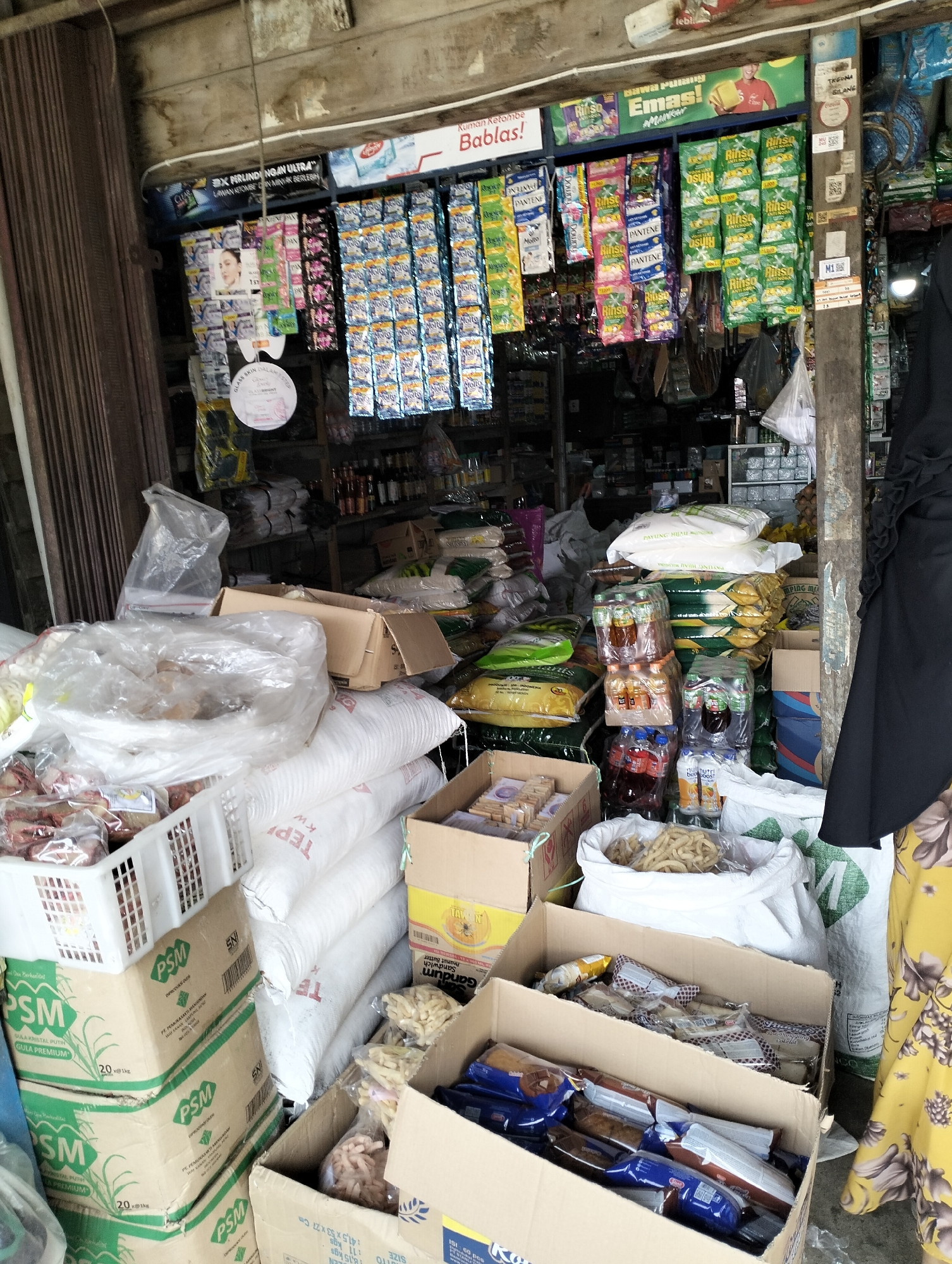 The image depicts the interior of a small, cluttered shop in Serdang Jaya, Indonesia, likely a general store or minimarket. The shop is packed with various goods, suggesting a busy retail environment. Products are displayed on shelves, hung from the ceiling, and stacked on the floor.

In the foreground, large white sacks, possibly containing rice or flour, are stacked high. In front of these sacks, there are several cardboard boxes, some open, filled with packaged food items like biscuits, snacks, and bottled drinks. One box is labeled "PSM GULA KRISTAL PUTIH GULA PREMIUM," indicating it contains premium white crystal sugar. Another white plastic basket holds more packaged goods. A large white plastic bag contains a pile of light-colored, irregular-shaped snack items.

Towards the left side of the image, a dark brown accordion-style door is partially visible. Above the shelves, banners and advertisements are hung, featuring product branding and promotional messages in Indonesian. Some of the visible brands include "RINSO," "PANTENE," and "Molto." A sign with the word "Babas!" is prominently displayed.

In the background, shelves are filled with a diverse array of packaged goods, bottles, and other items. A person, wearing a dark hijab and a yellow floral patterned garment, is partially visible on the right side of the frame, suggesting they are either a customer or the shopkeeper. The overall impression is one of a densely stocked, functional retail space catering to everyday needs in a local Indonesian community.