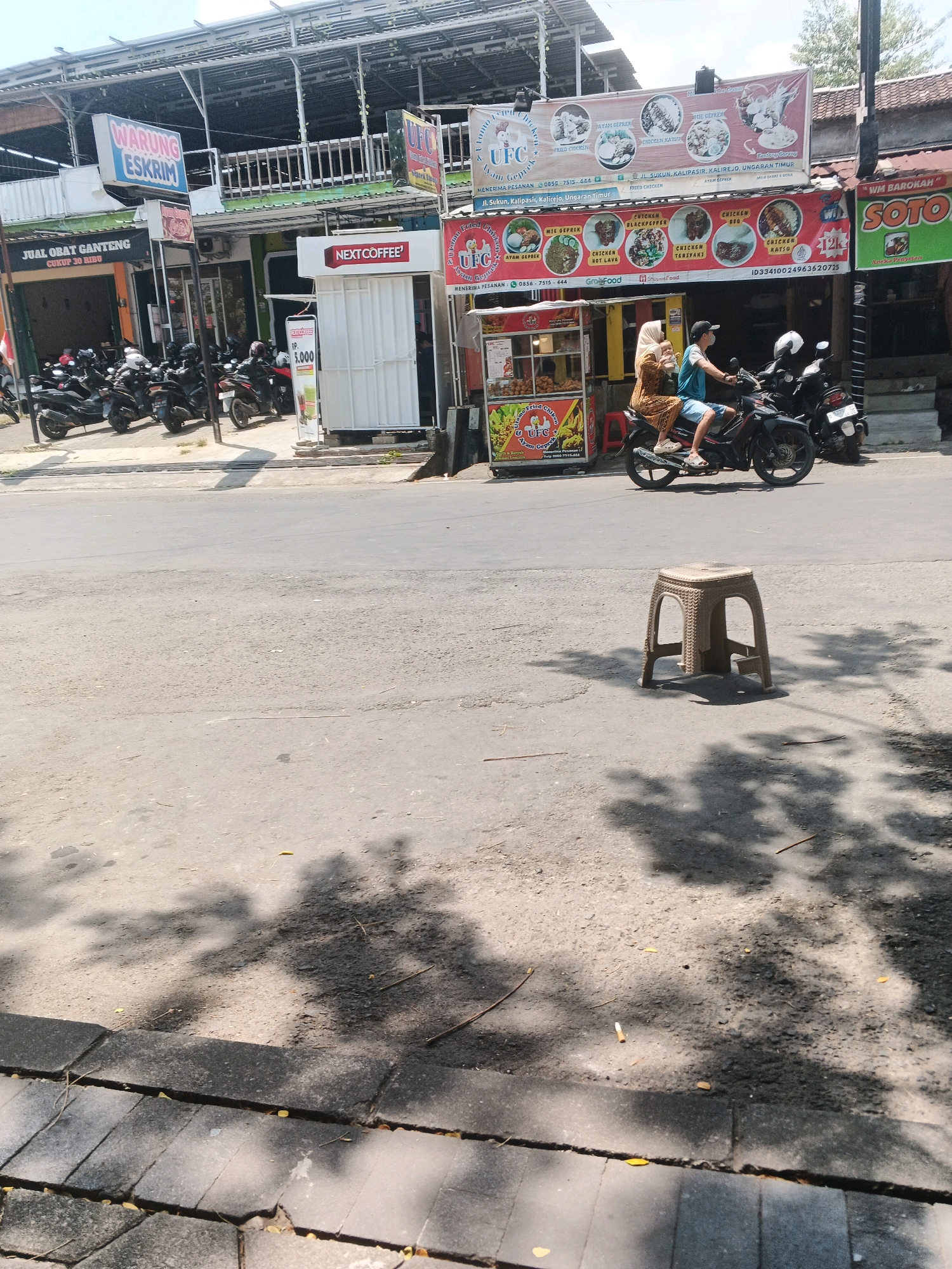In the city of Kalirejo, Indonesia, a street scene unfolds. The foreground is dominated by an asphalt road, with a textured, gray sidewalk to the lower left, marked by visible cracks. Sunlight casts dappled shadows across the road and sidewalk, suggesting it is daytime. A light brown plastic stool stands on the asphalt, centered in the mid-ground.
The background reveals a bustling street with several businesses. On the left, a sign for "WARUNG ESKRIM" (Ice Cream Shop) is prominent above a structure that appears to be a shopfront. Adjacent to it, a sign reads "JUAL OBAT GANTENG" (Sell Handsome Medicine). Numerous motorcycles are parked in front of these establishments. A white kiosk-like structure with a "NEXT COFFEE" sign is also visible.
Further to the right, a large banner displays images of food, advertising "UFC AYAM GEPREK" (UFC Fried Chicken). A food cart is positioned below this banner, with a person in a blue shirt and a woman in a brown headscarf and dress riding a motorcycle, heading towards the right of the frame. Another sign for "WM BAROKAH SOTO" (Barokah Soto Restaurant) is present on the far right. The buildings lining the street are mostly single or two-story structures with open-air sections and metal roofing. The overall environment suggests a commercial area in a tropical climate.