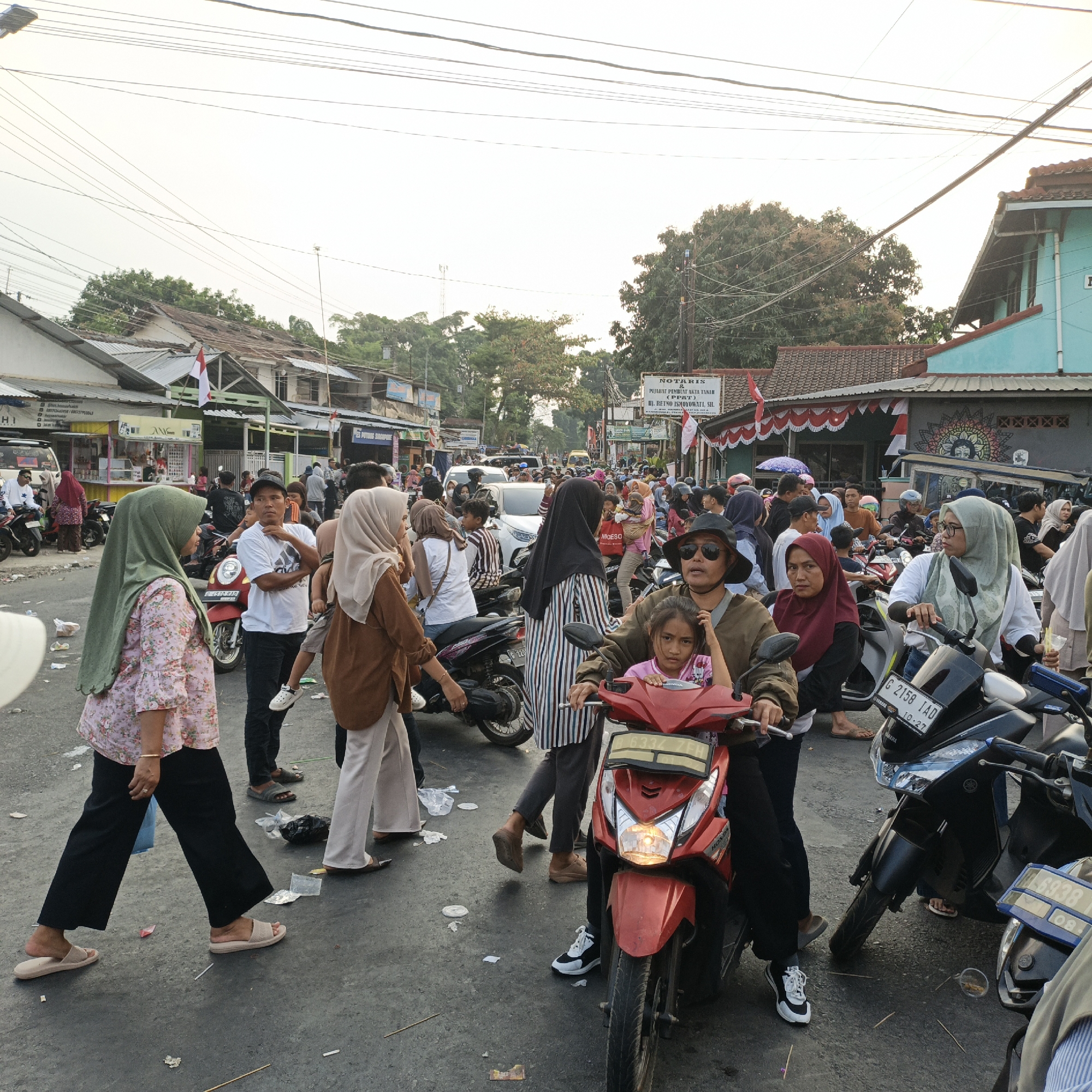 The image depicts a busy street scene in Randudongkal, Indonesia, likely during a daytime event or market. The foreground features several people on and around motorcycles. A woman in a green hijab and a floral-patterned shirt walks to the left, wearing black pants and beige sandals. Beside her, a man in a white t-shirt and black pants stands with his arms crossed. In front of them, a young girl sits on a red motorcycle, looking towards the camera. To the right, a man wearing a hat and sunglasses is seated on the red motorcycle.
Further back, a crowd of people are milling about, many also on motorcycles or walking. There are several buildings lining the street, with awnings and shops visible. Signs are present, including one for "NOTARIS & PEJABAT PEMBUAT AKTA TANAH". Indonesian flags are visible on some buildings. The street itself appears to be paved and shows some litter. The atmosphere suggests a lively, community gathering. The sky is overcast with visible power lines crisscrossing the frame.