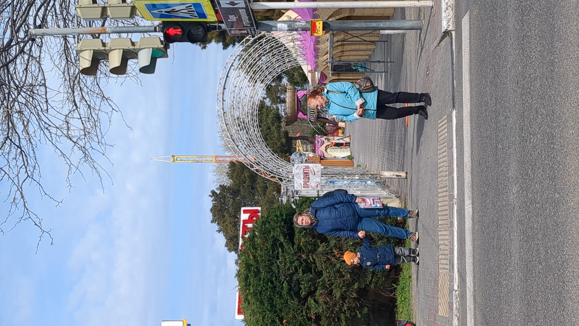 The image shows an outdoor scene on a bright, sunny day in Gelendzhik, Russia. In the foreground, a woman wearing a blue jacket and blue pants stands next to a young child in a dark blue jacket and an orange hat. They are positioned on a paved sidewalk, near a green bush and a red and white sign partially obscured by the foliage. To the right of the sidewalk is an asphalt road. Behind the woman and child, there is a circular metallic structure with a sign reading "ПРОДУКТЫ" (products). A tall amusement park ride is visible further in the background. Another woman, wearing a blue jacket and dark pants, is walking on the sidewalk in the background. Traffic lights are seen at the top of the frame, showing a red light. A street sign with a pedestrian crossing symbol is also present. The sky is blue with scattered clouds.
