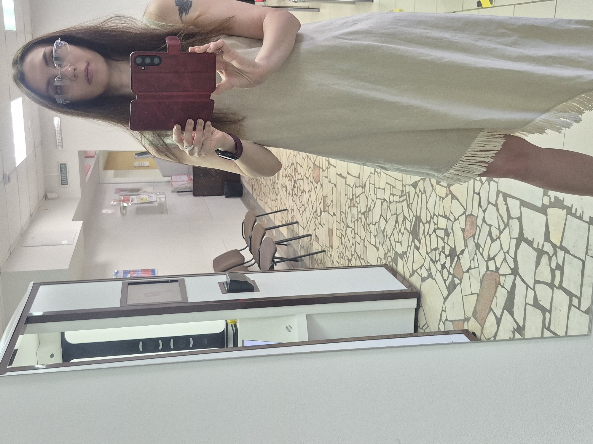 A full-length reflection in a mirror shows a woman in an unknown location within the city of Unknown, Unknown. The woman is holding a cell phone with a red case. She is wearing glasses and a light-colored dress with fringe at the hem. The floor visible in the mirror has a mosaic tile pattern. There are chairs and a white desk unit visible in the background, along with other features suggesting a public space.