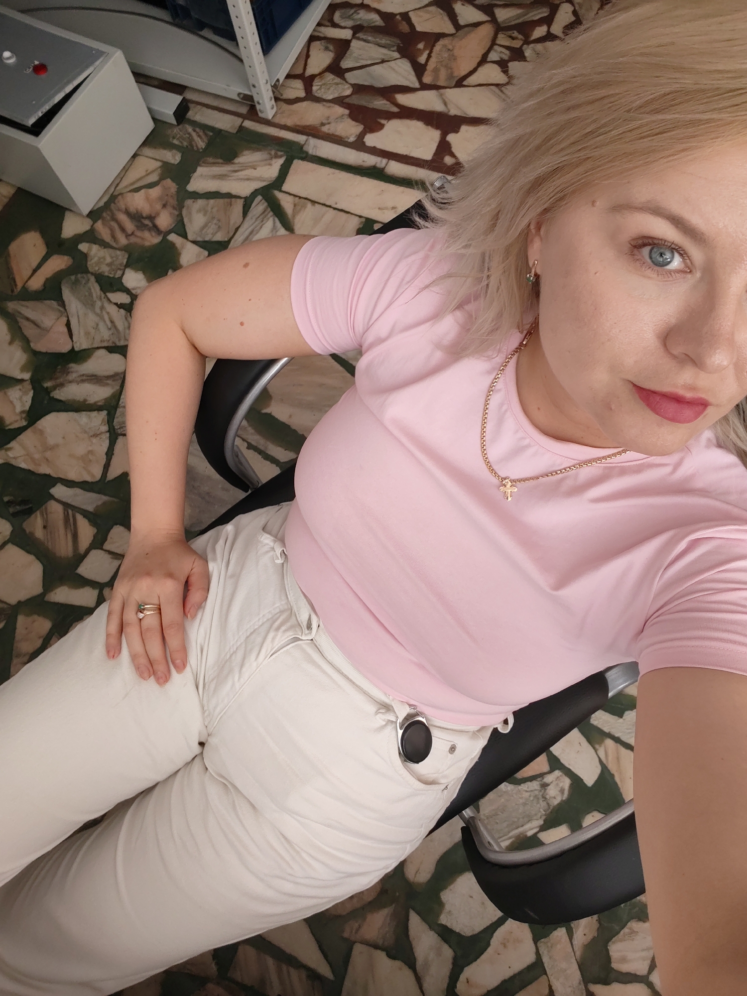 A woman with blonde hair and blue eyes is sitting in a chair, looking at the camera. She is wearing a pink t-shirt and white pants. A gold necklace with a cross pendant is visible around her neck, as well as a ring on her finger. The background shows a floor with a stone tile pattern, and a metal shelf unit is partially visible. The lighting suggests an indoor setting, possibly an office or a home. The woman has a neutral expression. There is no visible text in the image.