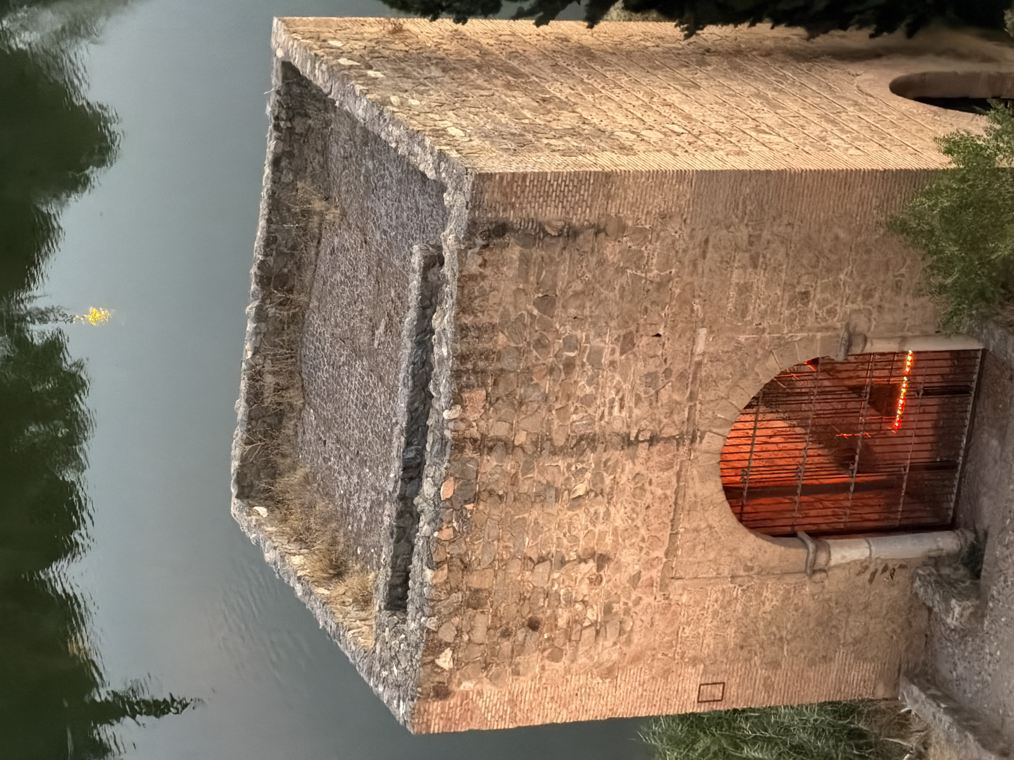 The image captures a stone tower with a cobblestone roof and a gate leading into it. The tower is situated next to a body of water, likely the Tagus River, as it is located in Toledo, Spain. The stonework of the tower is weathered and shows signs of age, with various types of stones and brickwork incorporated into its construction. The roof is covered in small, irregular cobblestones and sparse, dry vegetation grows along its edges. An arched entryway with a wrought-iron gate is visible on the front of the tower, and behind the gate, a warm orange light emanates from within. The water's surface reflects the surrounding environment, including hints of trees and possibly buildings. The scene appears to be illuminated by natural light, likely during twilight, given the soft glow and the muted colors. The tower is part of the historic landscape of Toledo, a city renowned for its medieval architecture and its strategic position along the Tagus River.