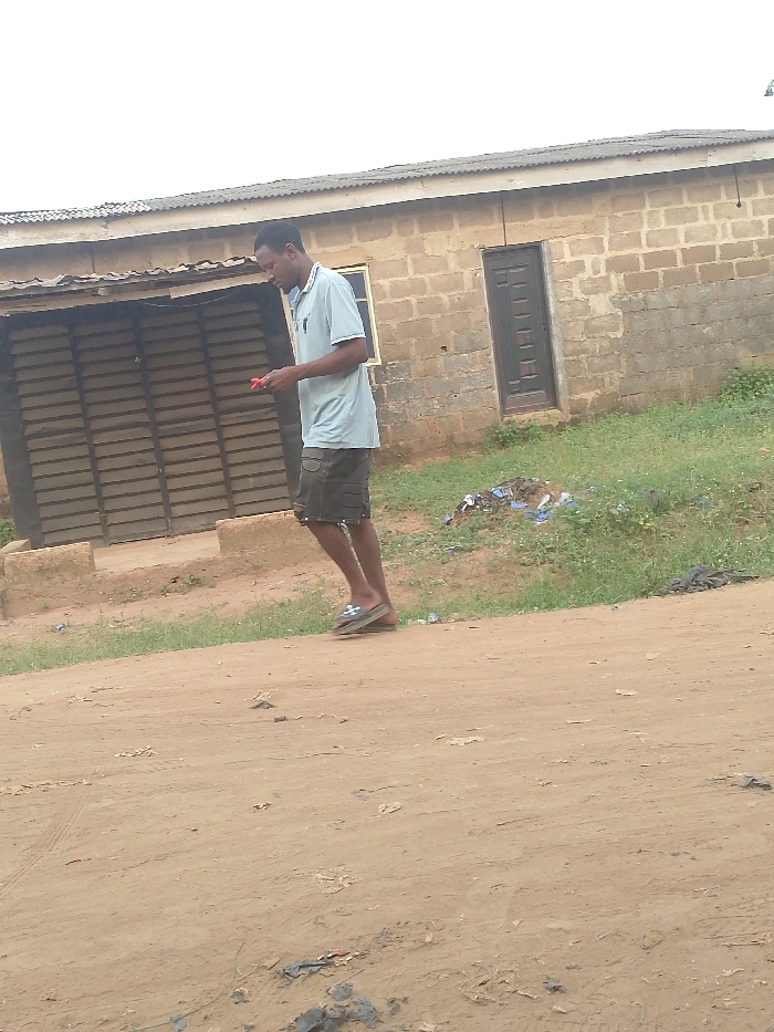 A man walks on a dirt road in front of a one-story brick building with a corrugated metal roof. The building has a dark door and a boarded-up opening. He wears a light-colored short-sleeved shirt, dark shorts, and flip-flops. He holds a small red object in his hand. There is some sparse vegetation and trash near the building. The sky is overcast.