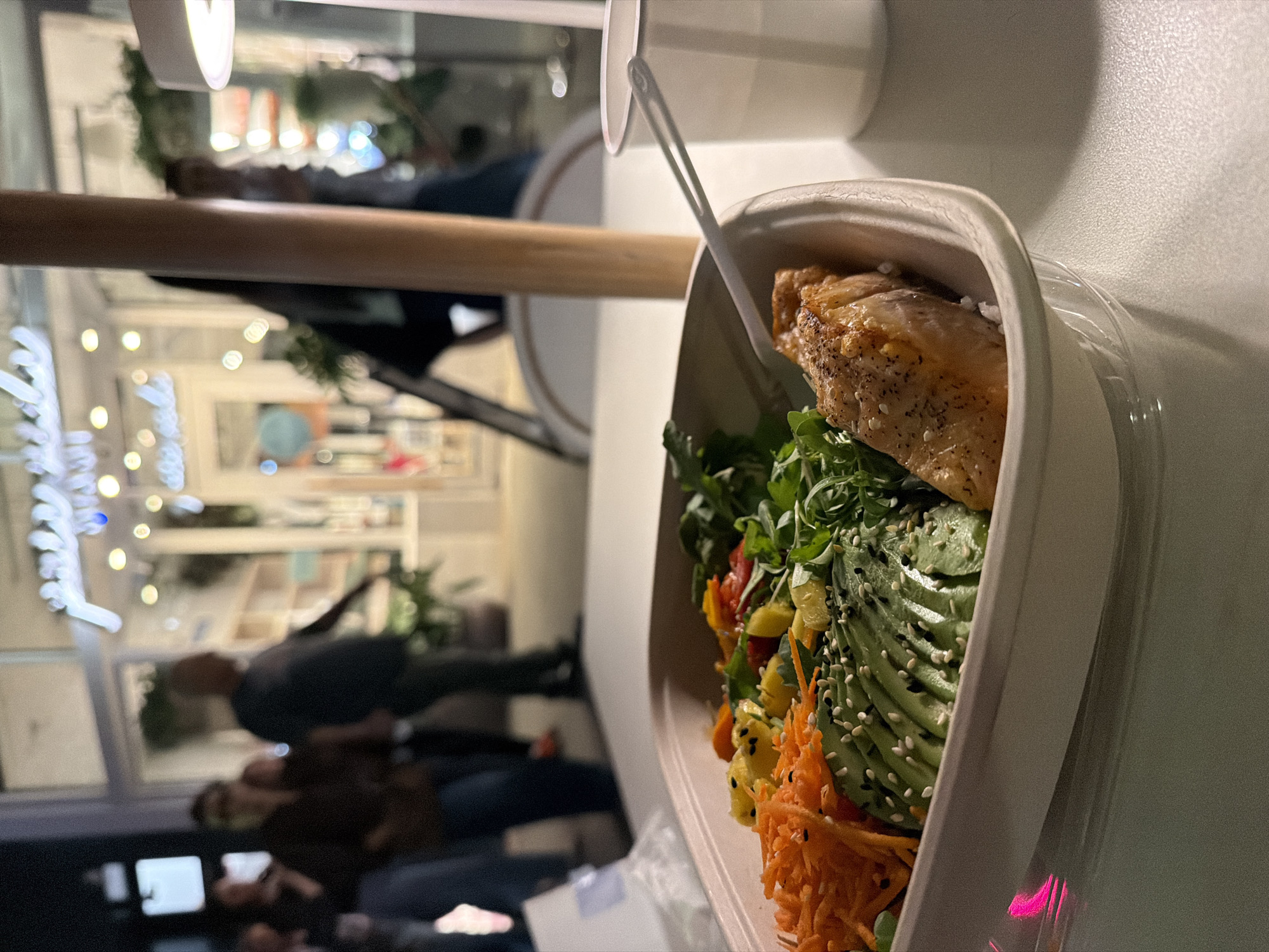 Here is a detailed description of the image:
**Scene Overview:**
The image shows a close-up of a food bowl containing what appears to be a salad with chicken, avocado, carrots, and other greens. A pair of chopsticks stick out from the bowl. The bowl is in a takeout container.
**Setting:**
The photograph appears to have been taken indoors, possibly at a cafe or restaurant, as the image shows a blurred background with people walking and other patrons visible behind a glass storefront. Given the presence of a storefront and people walking outside, this is likely in an urban setting.
**Subjects and Objects:**
* **Food Bowl:** The main subject is the bowl of salad. It includes components like cooked chicken, sliced avocado sprinkled with seeds, shredded carrots, and a mix of green leaves.
* **Takeout Container:** The food is contained within a light-colored, disposable container, which appears to have a clear plastic lid partially covering the contents.
* **Chopsticks:** A set of white chopsticks is seen protruding from the bowl.
* **Background:** The background is blurred, but it's evident that there are people walking, shop displays, and lights outside a glass window.
* **Drink Cup:** A white drink cup can be seen in the upper right of the image.
**Actions and Interactions:**
* **Eating/Preparing to Eat:** The presence of chopsticks indicates that the food is either being prepared to eat or has been partially consumed.
* **Urban Environment:** The blurred background suggests that the action is taking place in an urban environment.
**People:**
The people in the background are blurred due to the focus being on the bowl of food.
**Environment:**
The environment appears to be a modern urban cafe or restaurant setting.
**Location (Inferred):**
The location of the image is likely in New York City, United States of America, as that's the only location provided, and given the high population density and urban setting, that is a plausible place to find a trendy cafe/restaurant with takeout options.