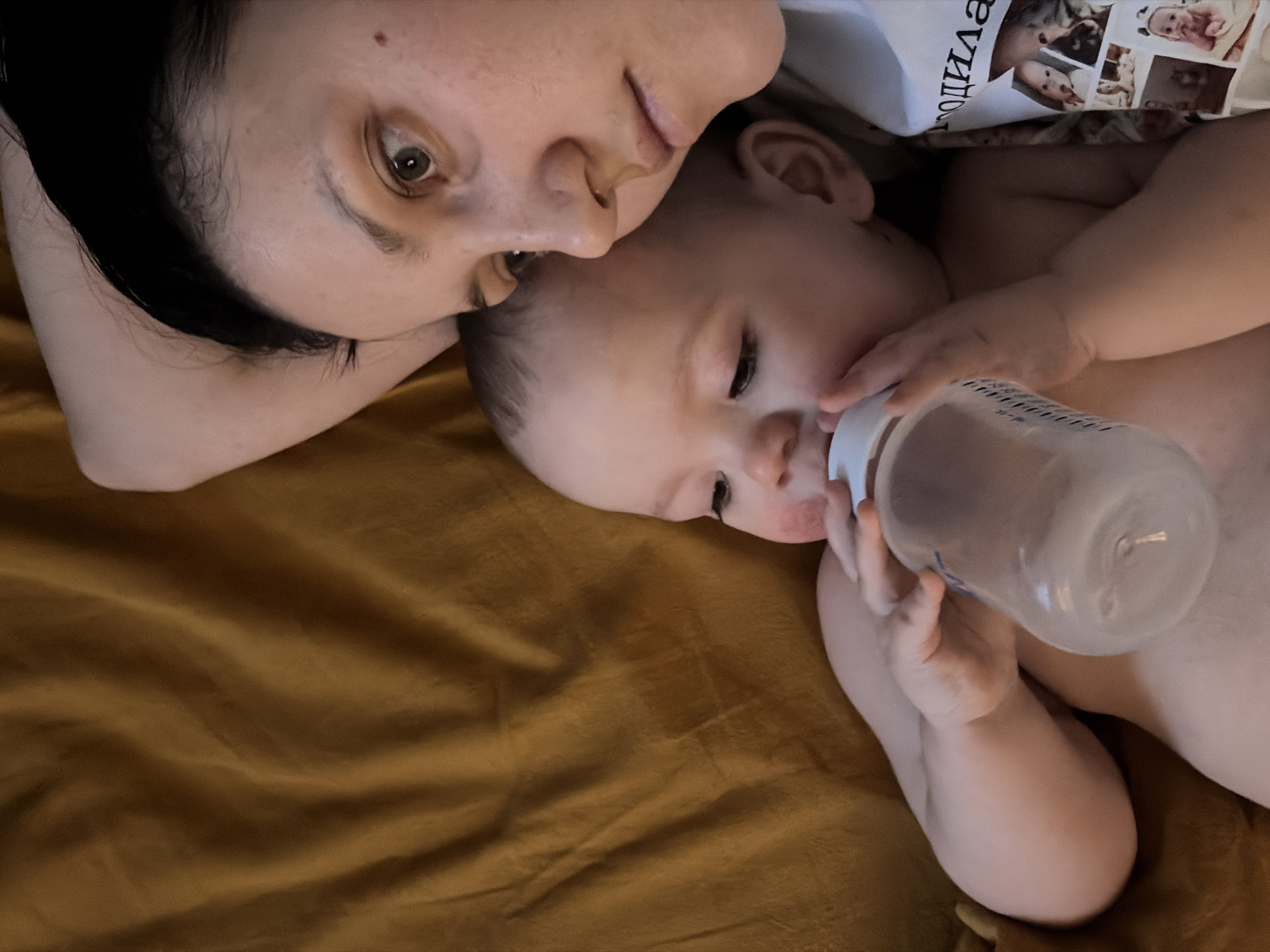A woman and a baby are lying down together on a golden-brown surface, possibly a blanket or a bedsheet. The woman is on her left side, with her head near the top-left of the frame. Her dark hair is pulled back, and she has fair skin. The baby is lying on its back and is holding a baby bottle with both hands, feeding from it. The baby has fair skin and is positioned to the right of the woman. The woman is wearing a white t-shirt with what looks like photos printed on it, along with some Russian text. The scene gives the impression of a tender moment between a mother and child. It's impossible to confirm the location in Chaikovsky, Russia, based solely on the visual information provided in the image.
