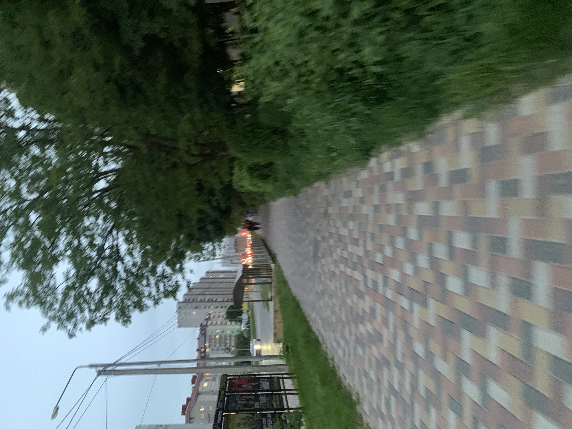 The image depicts a sidewalk in Stavropol, Russia. The sidewalk is paved with multicolored rectangular stones, and a strip of green grass runs alongside it. A row of trees lines the sidewalk on one side. Across the street, there are several tall apartment buildings. A street lamp stands near the edge of the sidewalk. In the distance, there appear to be cars stopped at a traffic light. A billboard stands near the street.