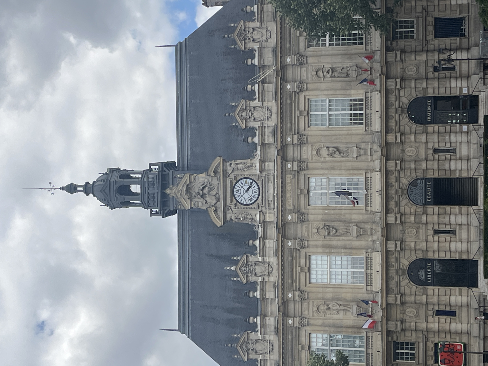This image shows the exterior of a large, ornate stone building, likely a town hall, in Ivry-sur-Seine, France. The building features a grand clock tower with a weather vane at its peak, intricately carved stonework, and numerous windows. Several French flags are visible, two of which are small and attached to flagpoles outside windows, while another appears to be displayed within a window.
The building's facade is adorned with sculptural elements, including busts and full-figure statues. The word "FRATERNITE" is inscribed above an arched entryway, and "EGALITE" and "LIBERTE" are visible above other arched doorways. The building's roof is made of dark, sloped tiles.
The sky is cloudy, suggesting it is daytime, but the specific time of day is difficult to determine precisely due to the angle and lighting. The weather appears to be overcast. The overall impression is one of historical architecture and civic pride, reflected in the grand design of the building and the national symbols displayed.