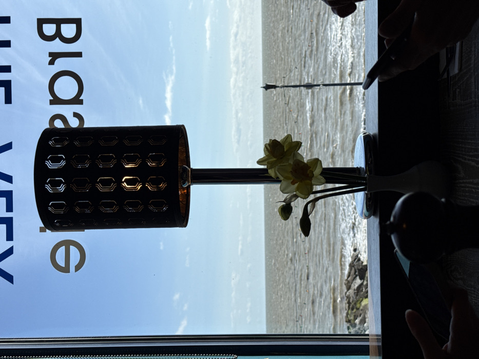 The image depicts an interior view from a restaurant or brasserie identified by the reversed window text "THE VEEX Brassiere" (likely "The View Brasserie"), overlooking the North Sea in Felixstowe, United Kingdom.
In the immediate foreground, a dark, textured table surface is visible. Two sets of silhouetted hands suggest the presence of at least two individuals. On the left, a hand holds a dark, rectangular object, while on the right, a hand grips a smartphone.
Central to the foreground is a dark, cylindrical table lamp with a chrome-colored stem and base. Its shade features a patterned cutout design, through which the illuminated bulb casts a warm light. To the right of the lamp, a small white vase contains a bouquet of yellow daffodils, comprising three open blooms and two closed buds. A dark, ornate object, possibly a condiment shaker, is partially visible on the table to the left.
Beyond the window, a light blue sky with scattered white clouds transitions to the expansive, choppy, grey-brown waters of the North Sea. The horizon line is clearly visible. To the left, a rocky breakwater extends into the water. In the mid-distance, several small, red/orange buoys are dispersed across the water's surface, and a thin, vertical pole structure, resembling a distant lamp post or pier element, rises from the sea towards the right. The exterior natural light is strong, backlighting and silhouetting the interior foreground elements.