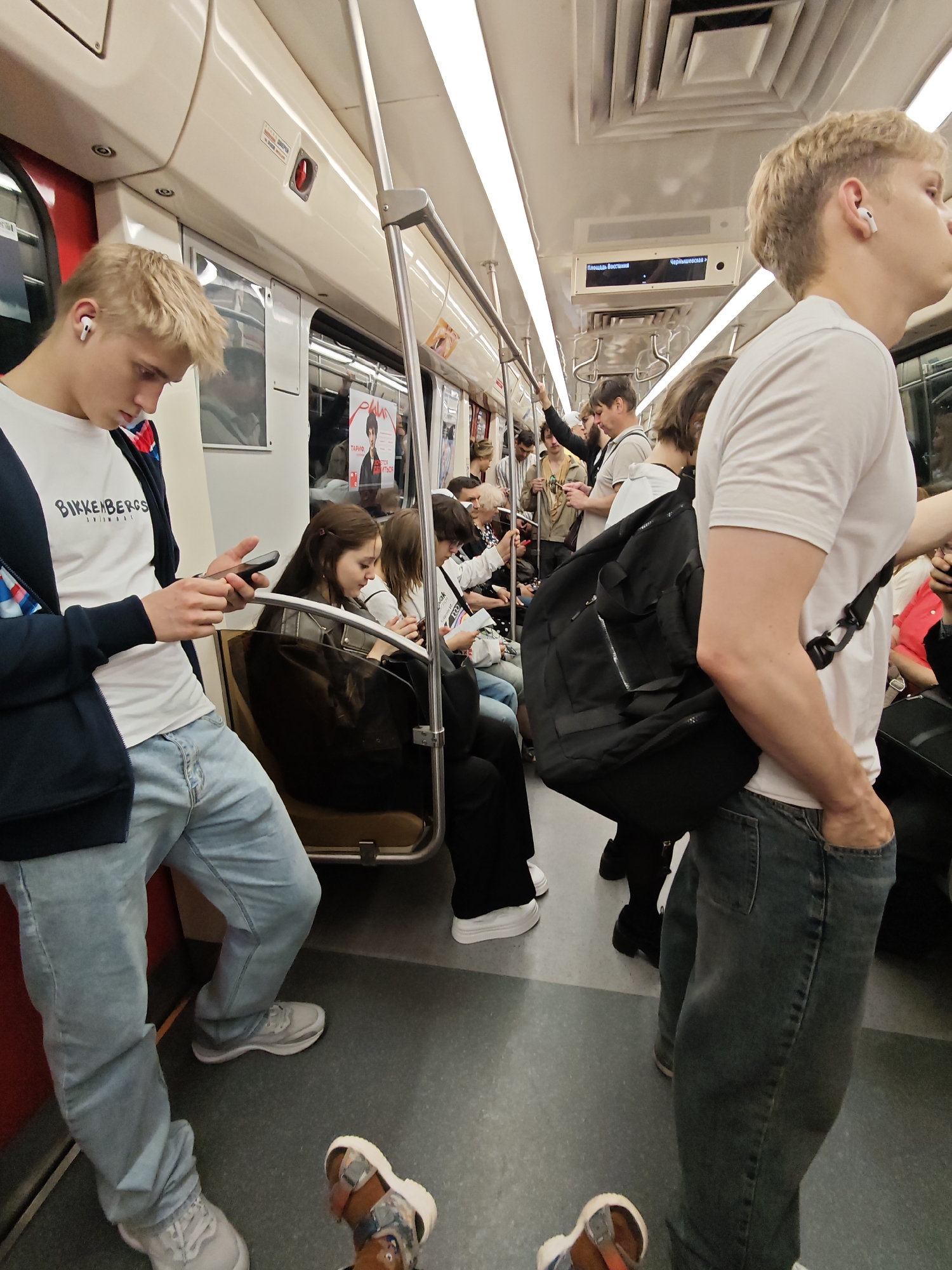 The image depicts the interior of a Saint Petersburg Metro train car. In the foreground, a young Caucasian male with blonde hair is standing and looking down at his smartphone, which he is holding in his hands. He is wearing a white t-shirt with the "Bikkenbergs" logo, a dark jacket with blue and red accents on the collar, and light blue jeans. He also has white wireless earbuds in his ears. To his right, a young Caucasian male with blonde hair is facing away from the camera, wearing a white t-shirt and dark jeans, with a large black backpack slung over his shoulder. His hand is in his front jeans pocket. The train car is crowded with other passengers, some of whom are seated on brown upholstered benches and others are standing. The train car features fluorescent lighting, metal handrails, and digital display screens above the windows showing station names, in Russian, such as "Площадь Восстания" (Ploshchad Vosstaniya) and "Чернышевская" (Chernyshevskaya). Advertisements are also visible on the train car walls. The overall environment suggests a typical rush hour commute on public transportation in Saint Petersburg.