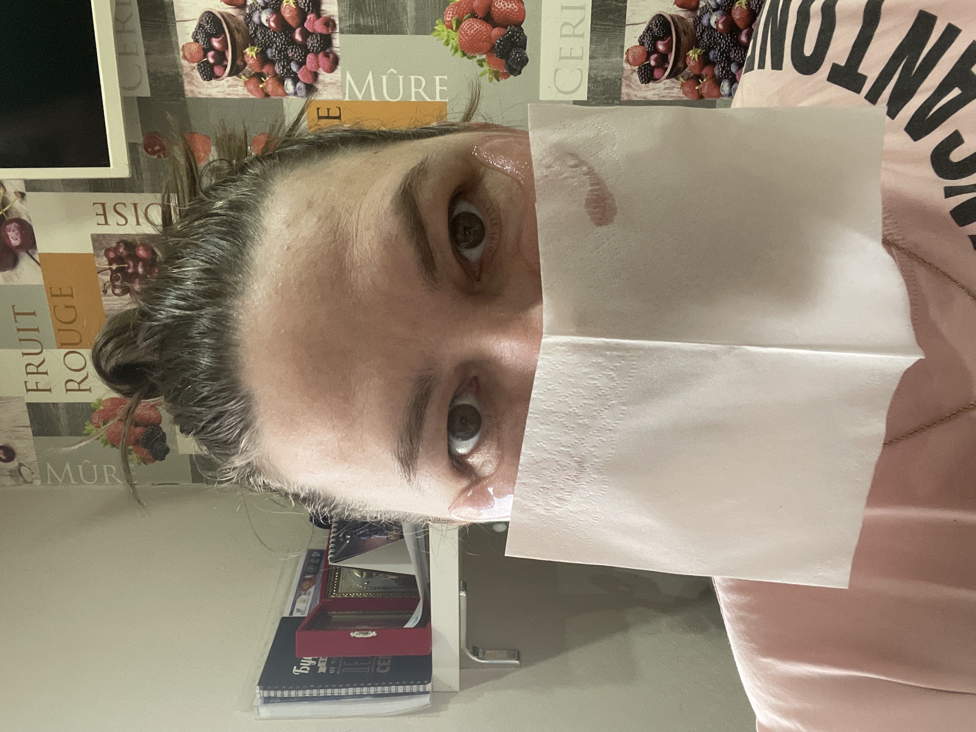 A woman with brown eyes and graying hair in a bun holds a white tissue against her face, obscuring her nose and mouth. A faint lipstick mark is visible on the tissue. The woman wears a light pink shirt with the partial word "VACATION" in large black letters. The background includes a wall with a fruit pattern and a bookshelf with books and a small decorative box. The location is unknown.