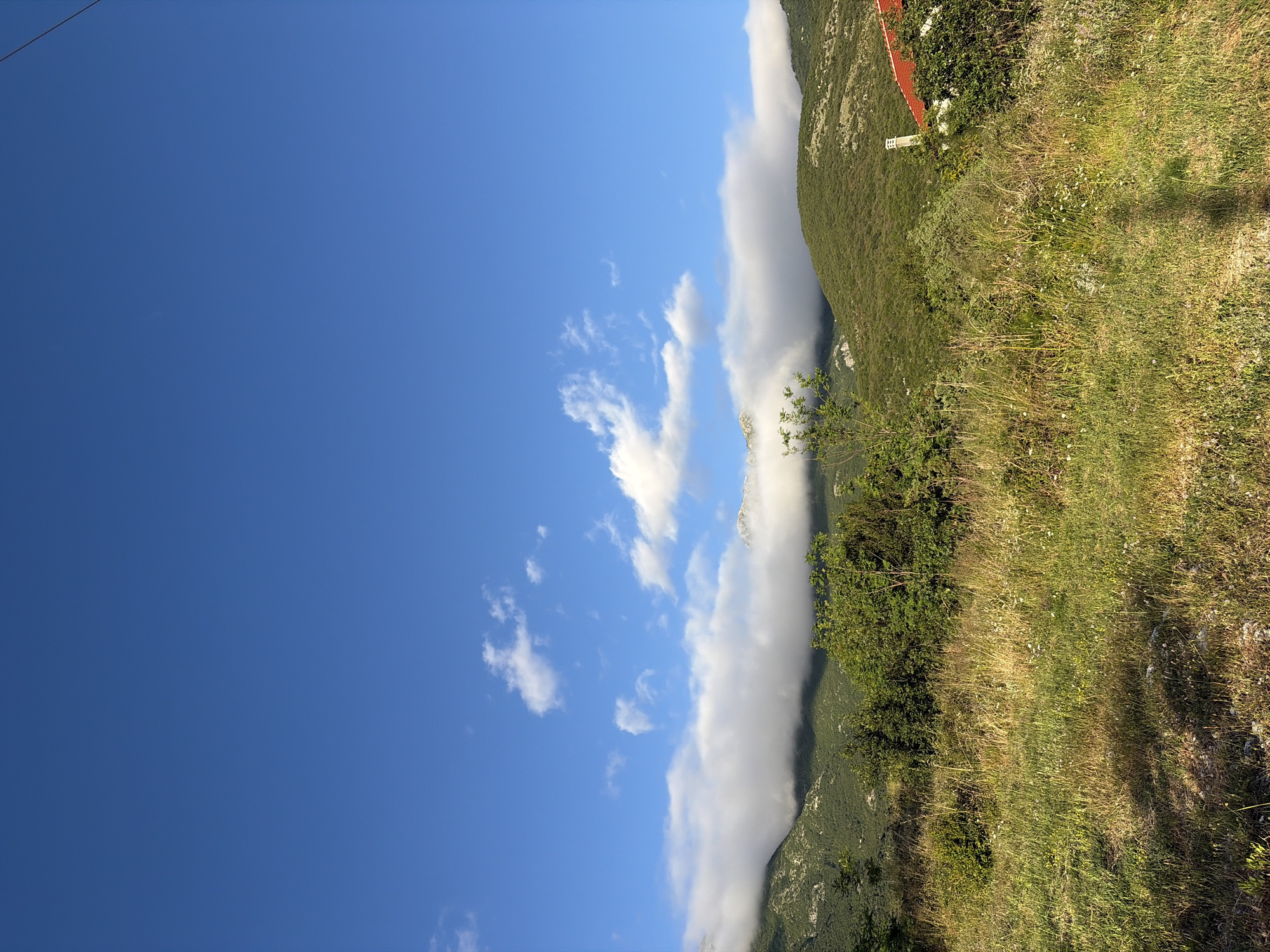 This image presents a serene mountain landscape under a bright, clear blue sky, situated in Bratešići, Montenegro. The left side of the frame is dominated by a vast expanse of deep blue sky, accented by several small, scattered white cumulus clouds.
A significant feature is a long, horizontal band of thick white clouds or fog that hugs the ridge of a large, verdant mountain, partially obscuring its upper slopes. This mountain, rich with dense green foliage, forms the right side of the composition. In the mid-ground, on the sloping mountainside, a small white building with a distinctive reddish-orange tiled roof and a chimney is visible, surrounded by lush greenery. Nearby, a small white sign is present, but any text on it is illegible.
The foreground consists of a sunlit grassy slope filled with various shades of green, dotted with small white and yellow wildflowers. Scattered bushes and patches of taller grass are also present. The scene is illuminated by bright daylight, suggested by the clear sky and subtle shadows on the grass. No people or specific activities are depicted, emphasizing the natural beauty and tranquil atmosphere of the mountainous region. The weather appears fair, characterized by the clear sky and the striking cloud formation along the mountain ridge.