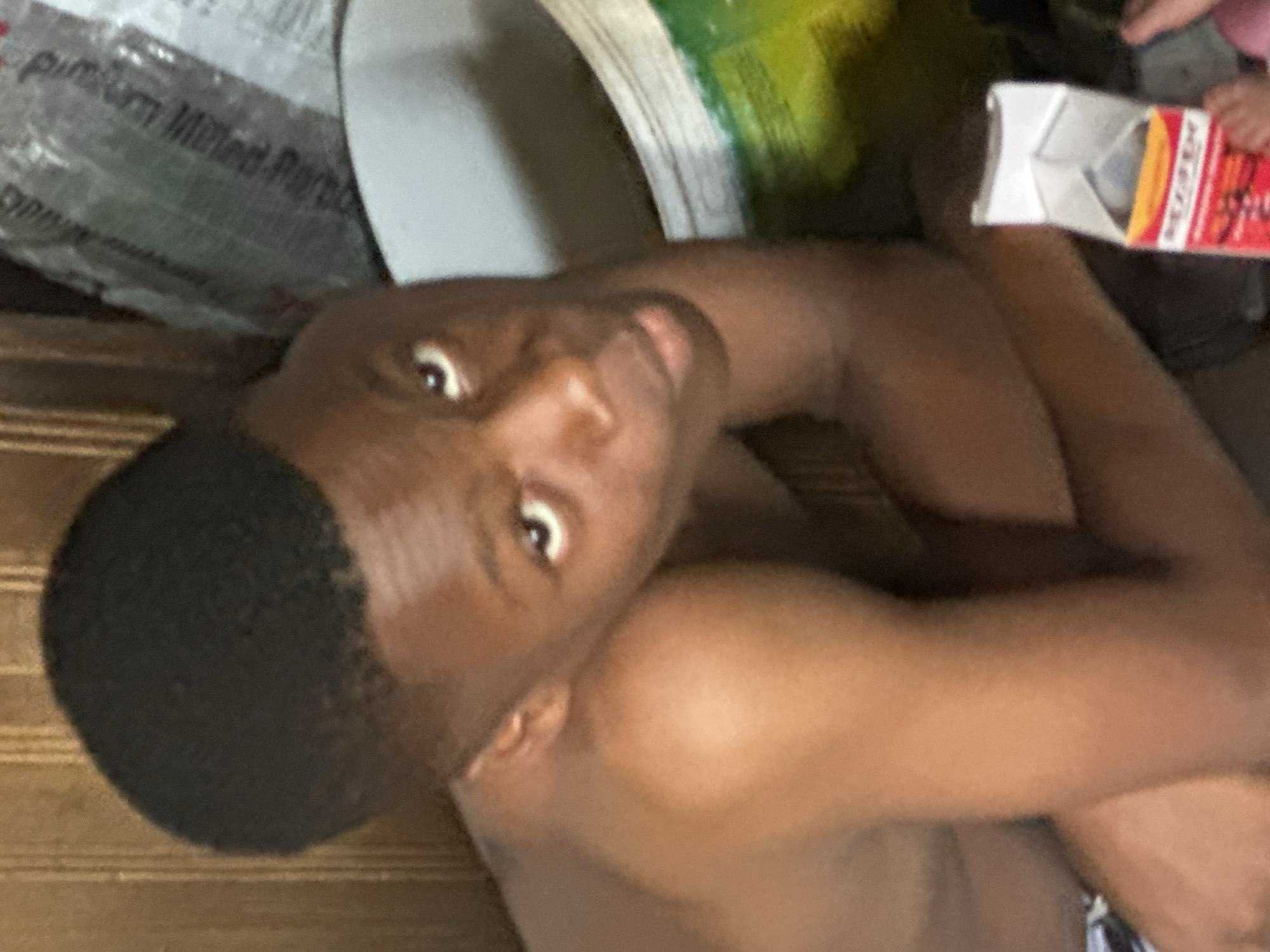 A close-up shot shows a dark-skinned young man with a short haircut looking directly at the camera. He appears to be shirtless and is situated indoors. Behind him are several objects, including a bag labeled "Pum Pum Milled" partially visible, and what seems to be a white container. To the right, a partially visible box in someone's hands displays red and orange colors. The scene is cluttered. There's no information to confirm this image was taken in the city of Aba, Nigeria.