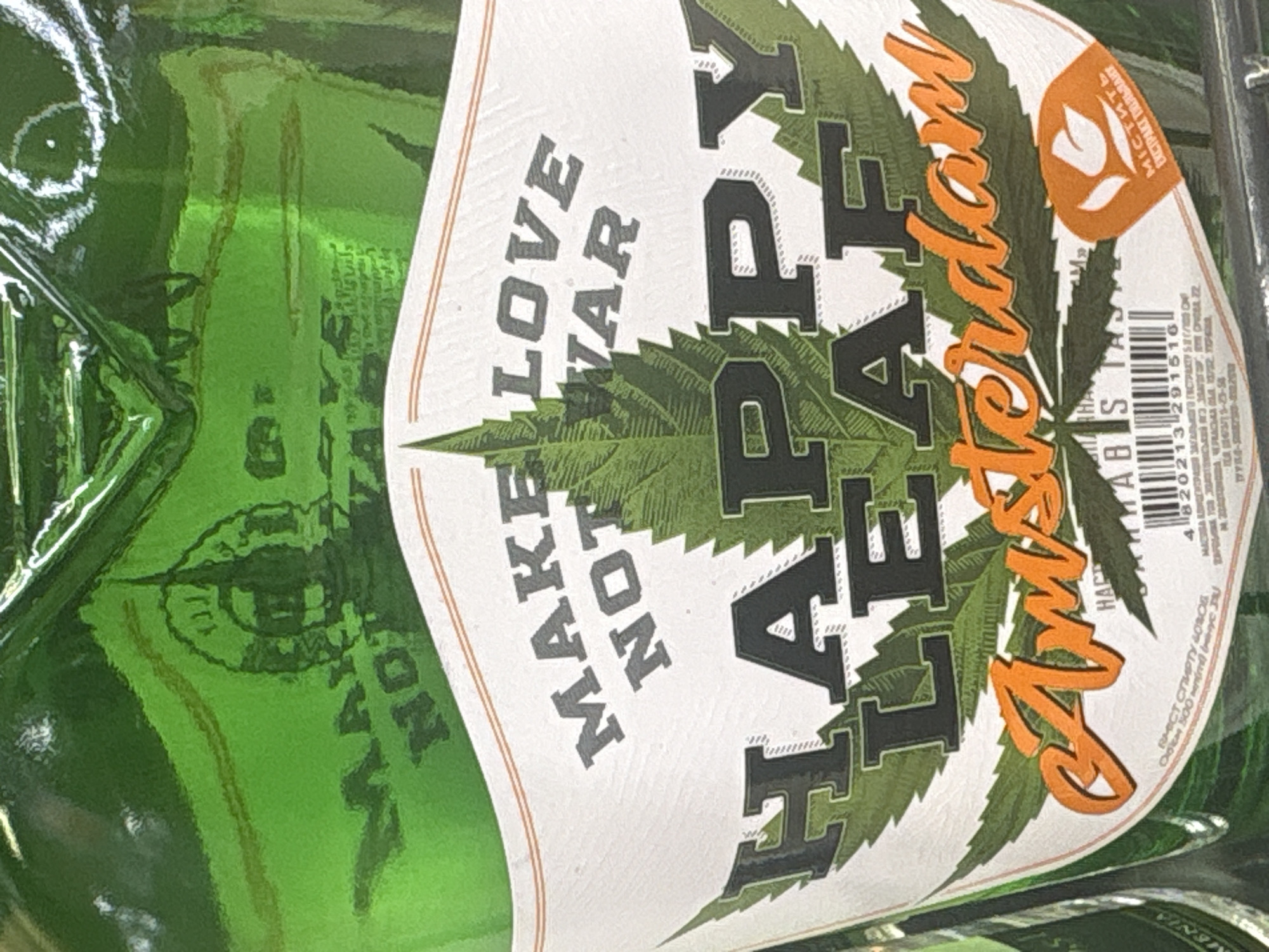 A green glass bottle with a white label is depicted in a close-up photograph taken in Yerevan, Armenia. The bottle's upper green glass surface, above the label, features a dark, subtly embossed cityscape silhouette, including a prominent spire. The white label is bordered by thin orange lines and centered with the grey uppercase slogan "MAKE LOVE NOT WAR." Below this, a large, stylized green cannabis leaf forms the background for the bold black uppercase text "HAPPY LEAF." Beneath "HAPPY LEAF," the word "Amsterdam" is written in orange cursive script. The lower right portion of the label displays an orange oval containing a white stylized tulip and the Ukrainian text "МІСТИТЬ ЕКСТРАКТ КОНОПЛІ" (Contains hemp extract). Adjacent to this, a barcode displays the number "4 820213 291516." Small Cyrillic text below the barcode indicates "ВМІСТ СПИРТУ 40% ОБ" (Alcohol content 40% vol) and "ОБ'ЄМ 500 мл" (Volume 500 ml), with partially visible English text identifying "CANNABIS TASTE." The bottle's surface reflects bright artificial light, and a portion of a similar green bottle is visible to the left. A dark horizontal surface, likely a retail shelf, is visible at the bottom of the frame.