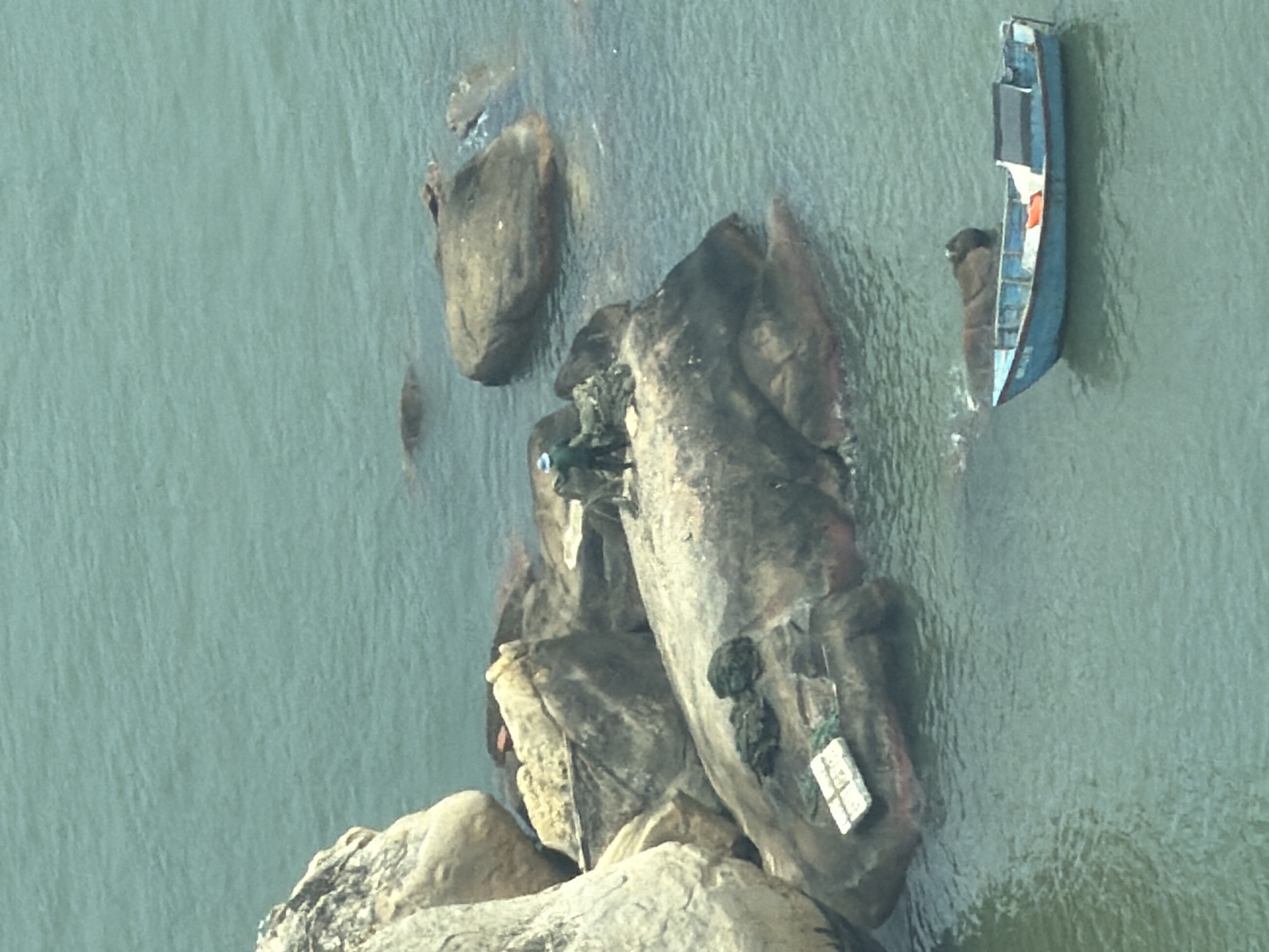 The image depicts a coastal scene near Nha Trang Ward, Vietnam. A small, blue boat is partially submerged in the water, adjacent to a cluster of large rocks. The water is a muted green. The rocks are varied in size and shape, with some showing signs of weathering and possible human interaction, such as small items attached to the rock surface. The angle of the shot is a top-down, high-angle view. There are no visible people in the image.