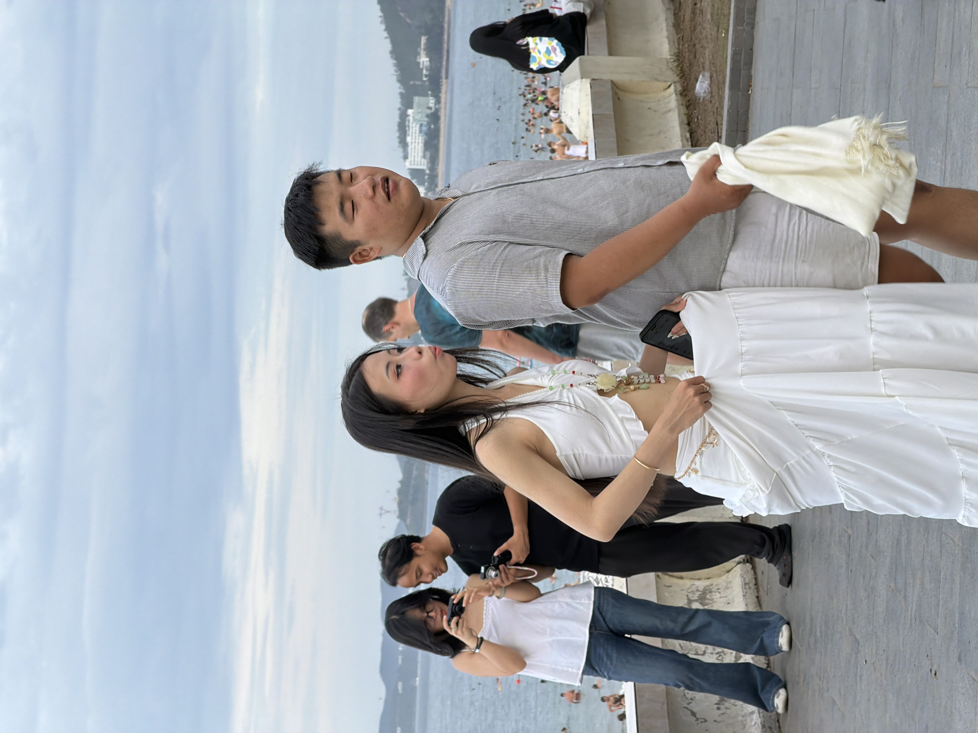 The image depicts a scene on a promenade in Nha Trang, Vietnam, overlooking the beach. In the foreground, a young Asian woman in a white halter-neck crop top and a tiered white maxi skirt is walking with a young Asian man in a light grey pinstriped short-sleeved shirt and white shorts. The woman holds a black smartphone in her left hand. The man is carrying a white, fringed scarf in his right hand. Behind them, to the left, another Asian woman in a white tank top and blue jeans is holding a black camera, and an Asian man in a black t-shirt and dark pants is holding a camera. Both appear to be taking photos. In the background, the sea is visible with numerous people swimming and sunbathing on the beach. The coastline is lined with buildings and hills. The sky is overcast with light grey clouds.