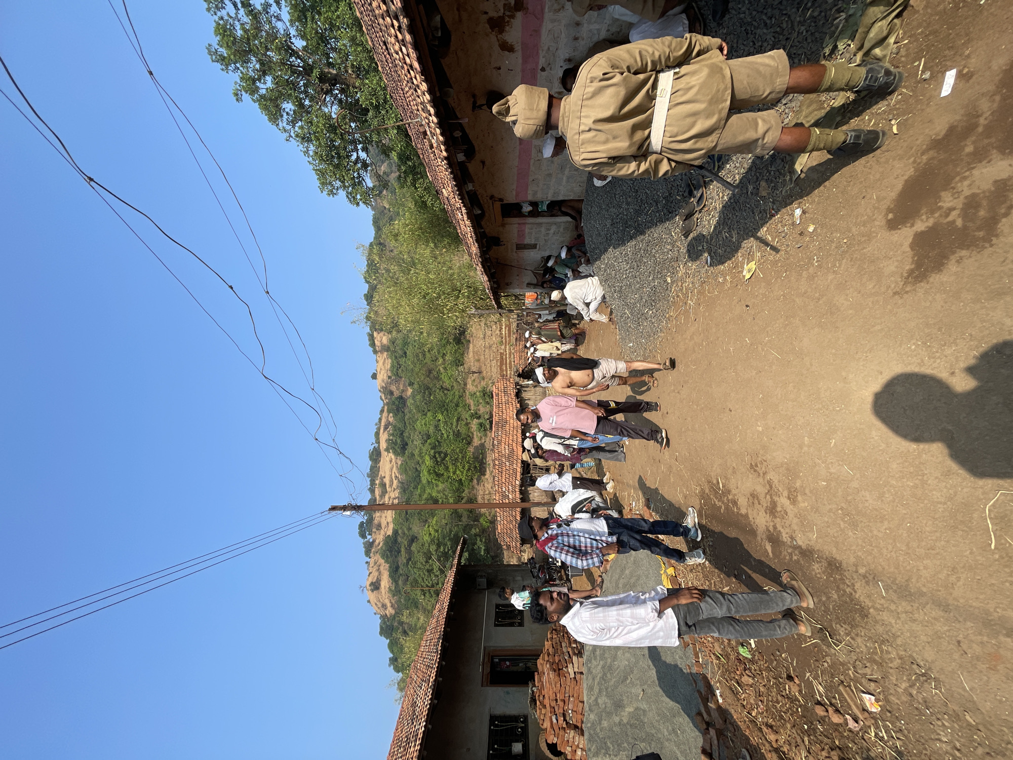The image depicts an outdoor scene in Sonurle, India. A line of people, possibly waiting or queuing, stretches along a dirt path adjacent to a building with a red-tiled roof. The building appears to be a simple structure, and there are stacks of bricks near it. Some people are wearing casual clothing, including shirts, pants, and some head coverings. One person is shirtless. At the head of the line stands a man in a tan uniform with a distinctive hat and boots. The scene is set against a backdrop of green hills and a clear blue sky. Power lines are visible overhead. The shadows suggest it is daytime.