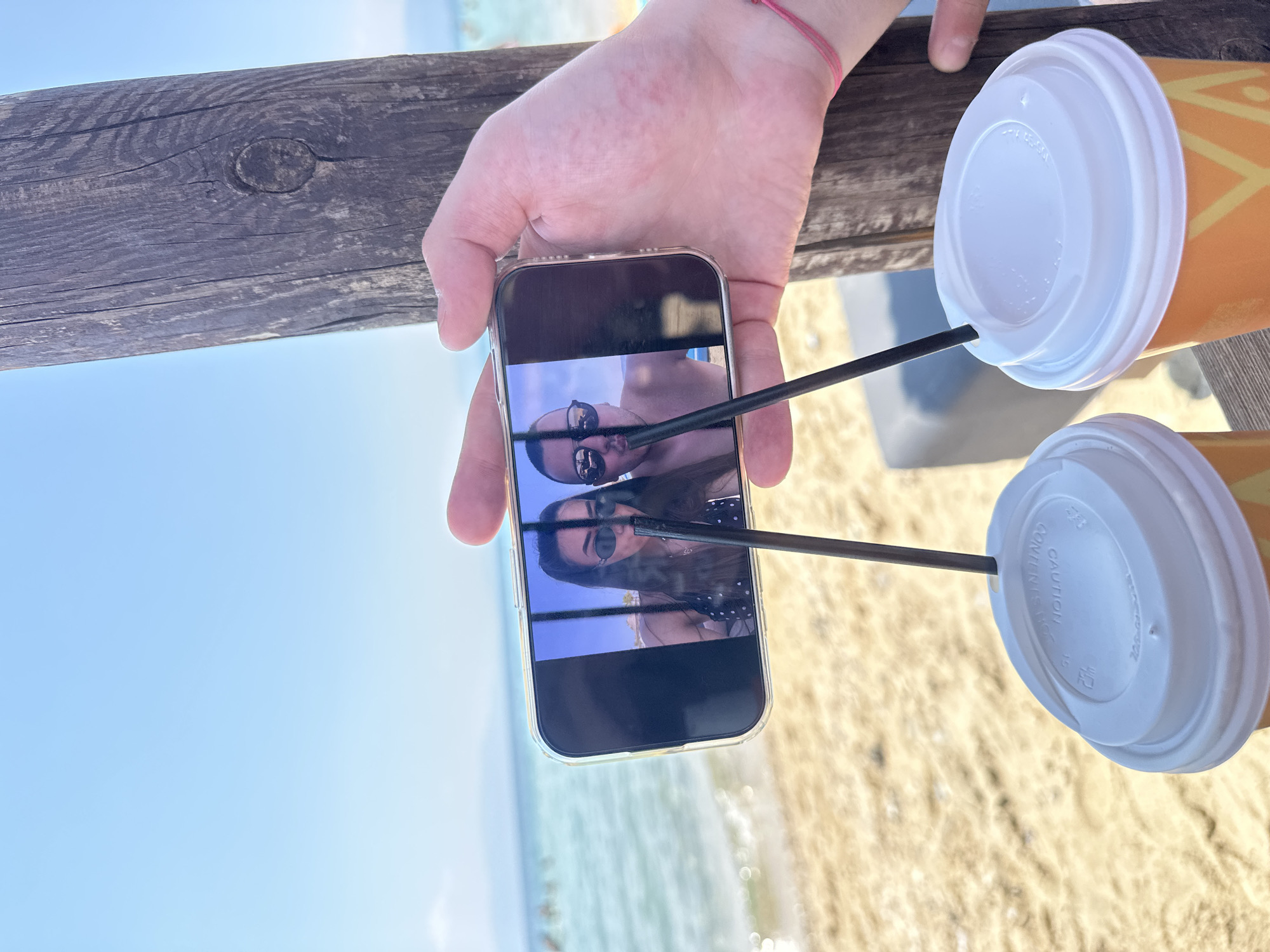 In the foreground, a person's hand holds a smartphone horizontally, displaying a selfie of two individuals. The smartphone is positioned in front of two disposable coffee cups with white lids and black straws. The background features a beach with sand, the sea, and a clear blue sky. A weathered wooden post is visible to the right of the hand and smartphone. The scene appears to be set at Ofrynio Beach, Greece.
