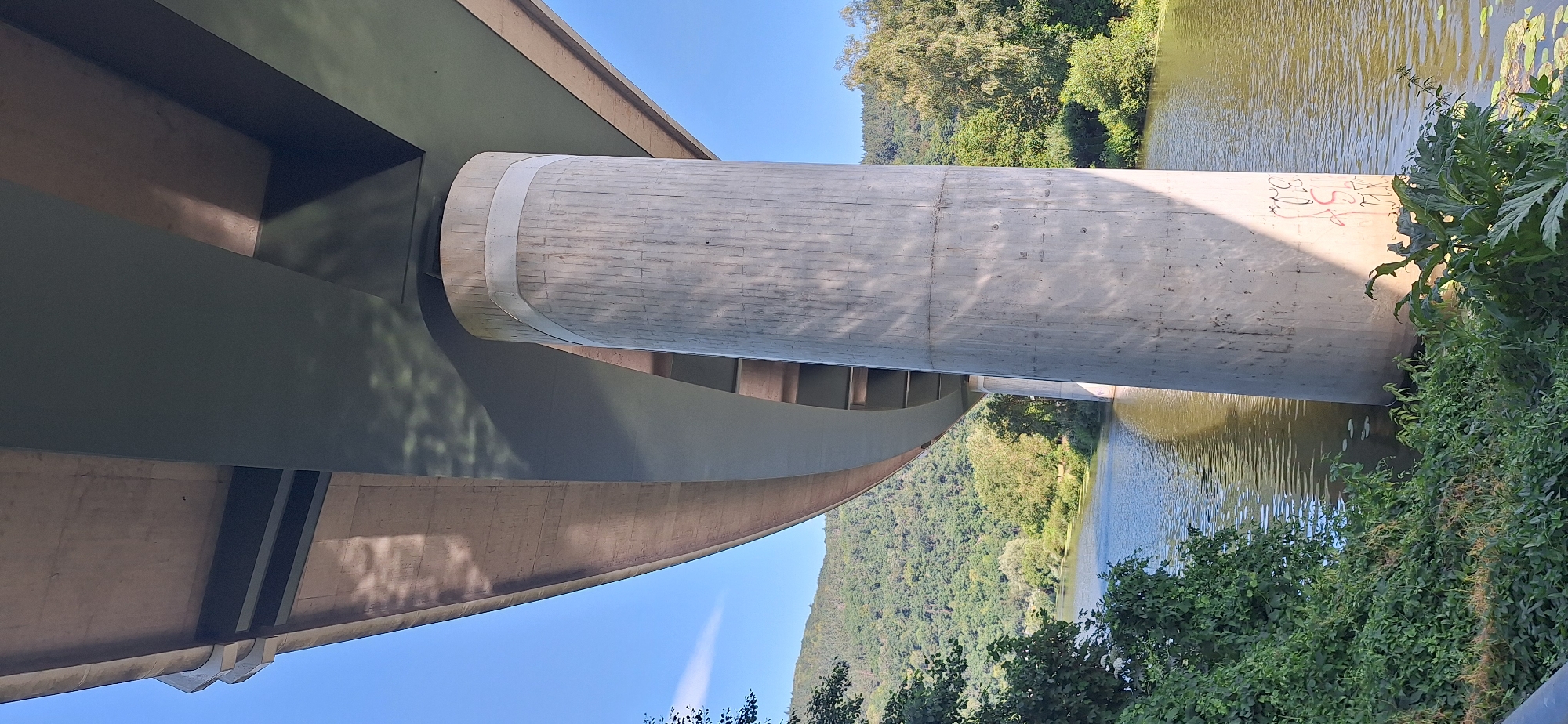 The image shows a concrete pillar supporting a bridge over a river in Fachbach, Germany. The pillar is cylindrical and textured with vertical lines. Two light-colored bands encircle the top of the pillar. Graffiti in red and black is visible on the lower part of the pillar. The river flows gently, with reflections of the bridge and trees on its surface. Lush green trees and vegetation cover the hilly landscape in the background. The sky is clear and blue.