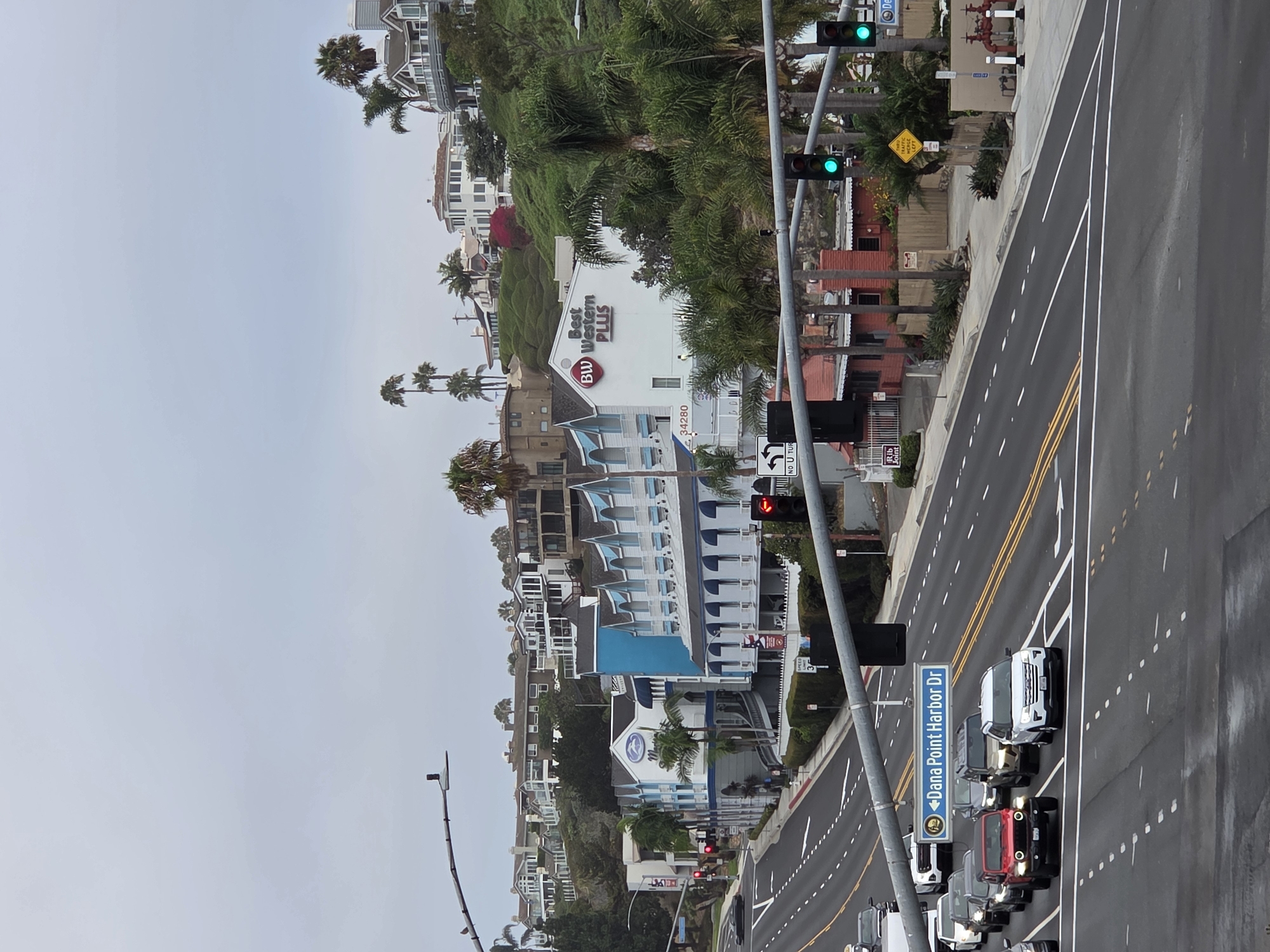 The image shows a street scene in Dana Point, California, USA, on an overcast day. The foreground features a multi-lane road with several cars stopped at a traffic light. A blue and white sign indicating "Dana Point Harbor Dr" is visible. Beyond the road, a hotel with blue and white architecture is situated on a hillside. The hotel has a sign that reads "Best Western PLUS". In the background, several houses and buildings are scattered across the hilly landscape, with palm trees dotting the scene. Traffic lights are visible, with some showing green. A speed limit sign indicating 35 is also present.