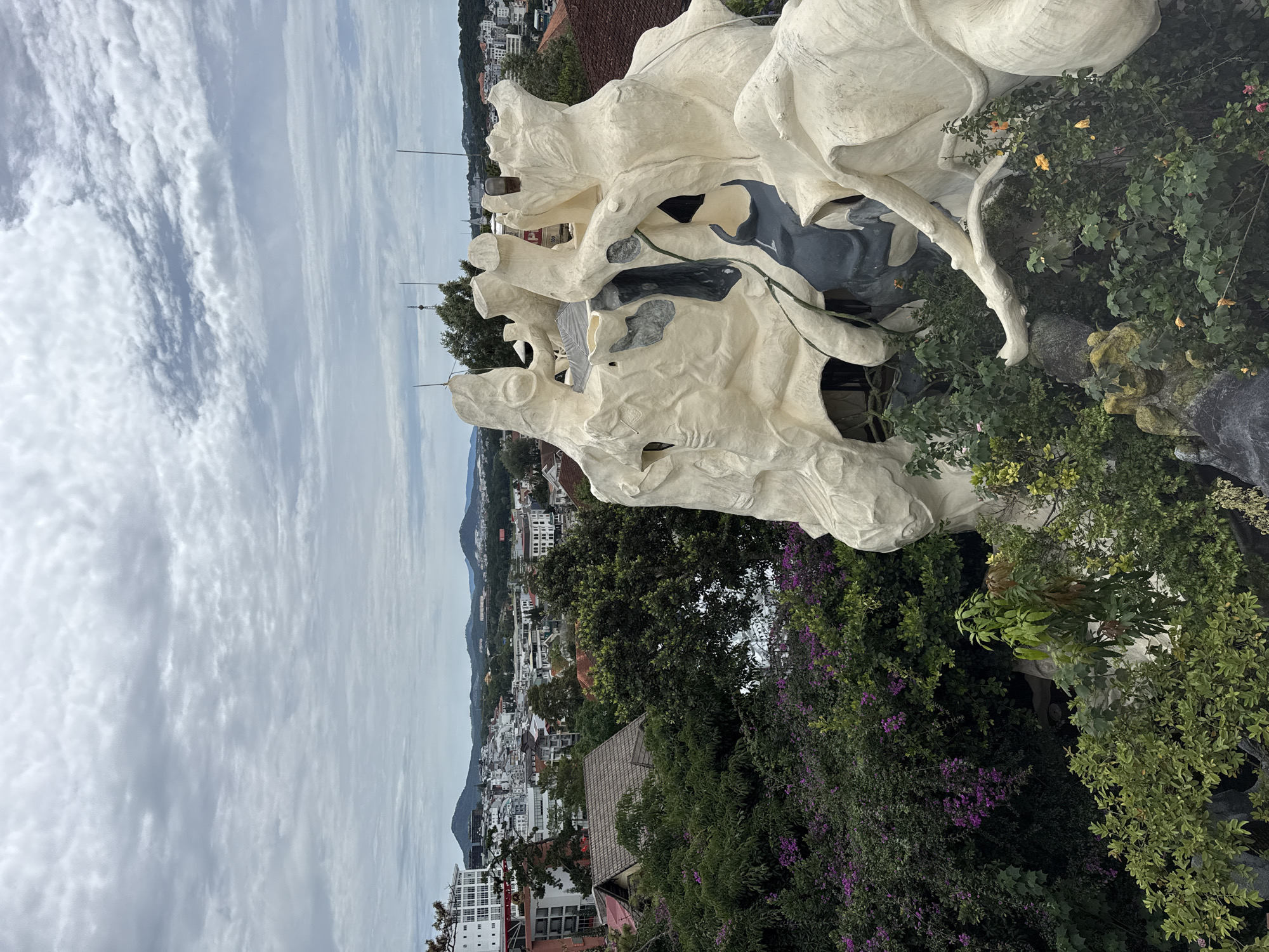 The image depicts a view from the Crazy House in Da Lat, Vietnam. The foreground is dominated by a large, abstract architectural structure made of light-colored, rough-textured material, resembling a tree or cave formation. This structure is interspersed with green foliage, including trees and flowering bushes with purple blossoms. In the midground, a dense urban landscape unfolds, comprising closely packed buildings of varying heights and styles, predominantly white and red-roofed, set against a backdrop of rolling hills. The sky above is overcast, with a layer of striated clouds. Several tall antennas are visible, puncturing the skyline. The image is slightly tilted to the left.