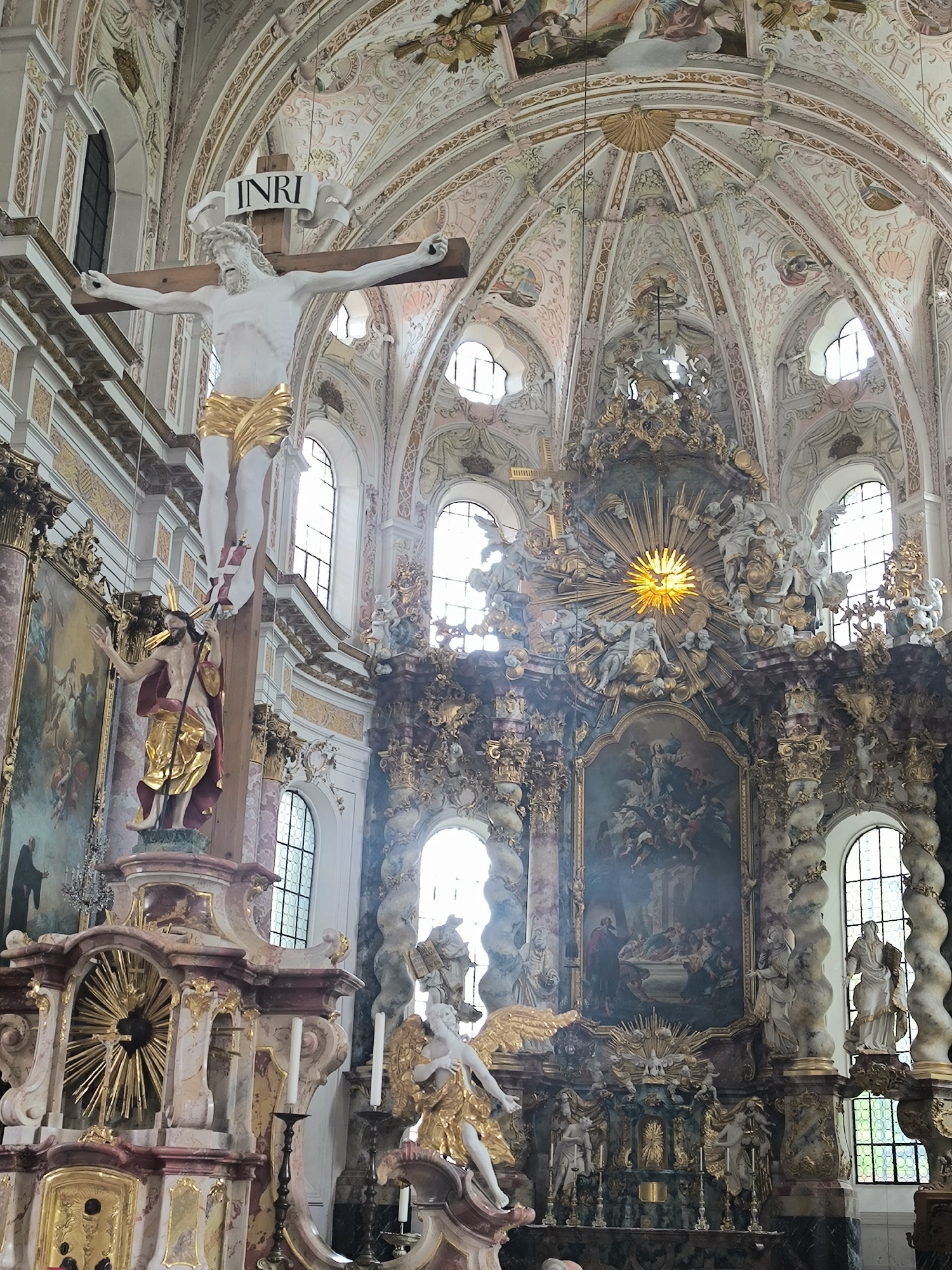 The image depicts the ornate interior of a church, specifically the interior of the Saint Korbinian’s Cathedral in Freising, Germany. The scene is filled with elaborate Baroque architecture, including painted vaulted ceilings, arched windows, and decorative moldings. The focal point of the left side is a large crucifix featuring a white figure of Christ. Below the crucifix is a golden statue of Christ. A grand altar stands on the right side. The altar is adorned with sculptures, paintings, and golden accents. Several statues, including angels, are seen throughout the scene. The architecture is highly detailed, suggesting a place of worship.