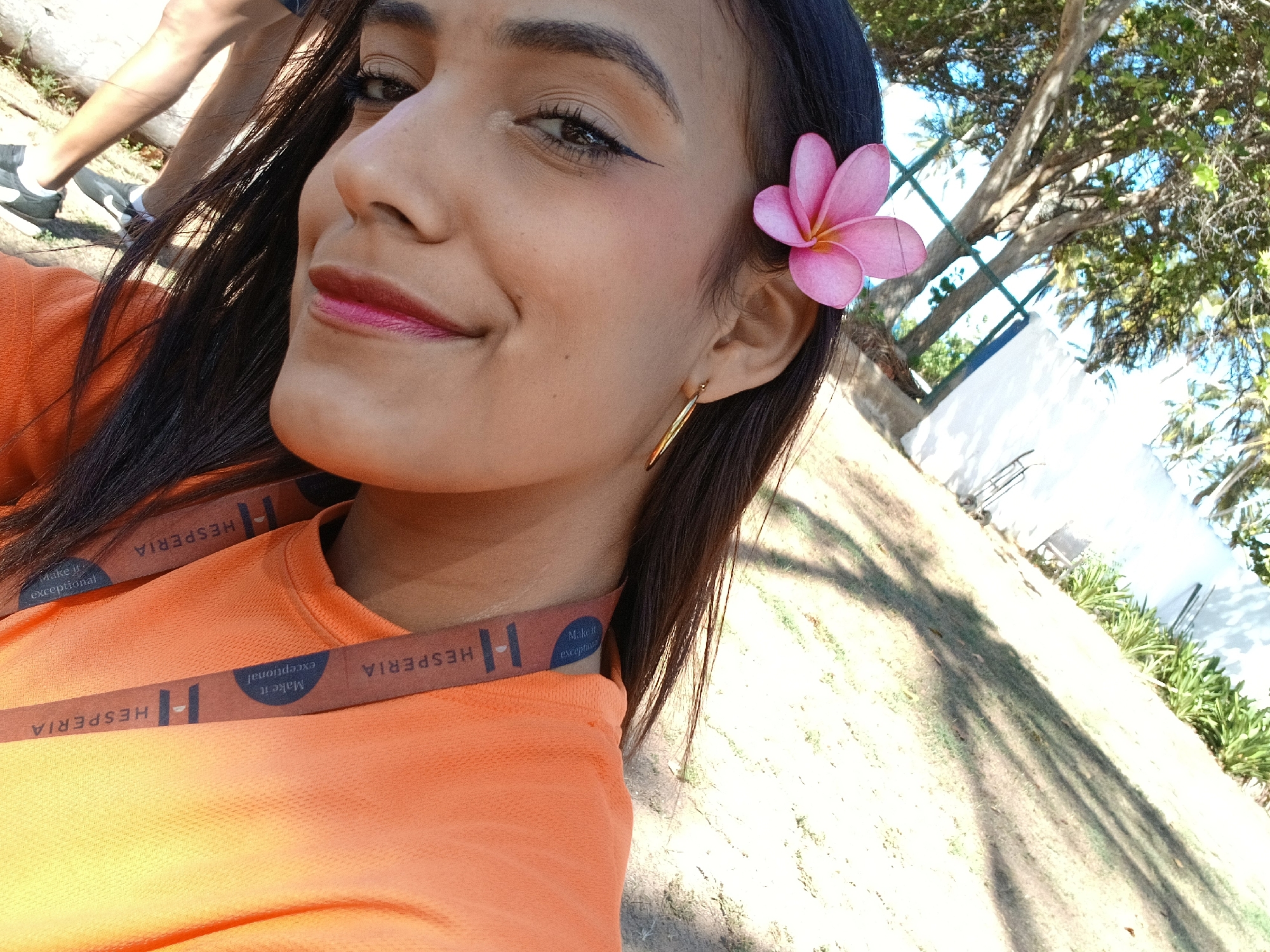 A close-up, eye-level selfie captures a young woman smiling outdoors on a bright, sunny day. She has dark, long hair, parted to her left, and wears a pink plumeria flower tucked behind her right ear. Her makeup includes black winged eyeliner and a two-toned lipstick, transitioning from a darker fuchsia on the inner part of her lips to a lighter pink on the outer edges. She wears a gold, elongated hoop earring in her right ear.
She is dressed in a vibrant orange, crew-neck t-shirt. A brown lanyard with white text is visible across her chest and shoulder. The text on the lanyard clearly reads "HESPERIA" repeated several times, along with the slogan "Make it exceptional" inside a blue oval graphic.
The background shows lush green trees and foliage under a bright sky, suggesting an outdoor, possibly tropical or resort-like setting. Strong shadows fall across the light-colored ground in the mid-ground and background, indicating direct sunlight, likely during the day. Part of another person's legs and shoes (dark sneakers with white soles) are visible in the upper left background, suggesting they are standing near her. The overall impression is one of warmth and happiness. The location context of Playa El Agua, Venezuela, aligns with the tropical flora and resort branding visible.