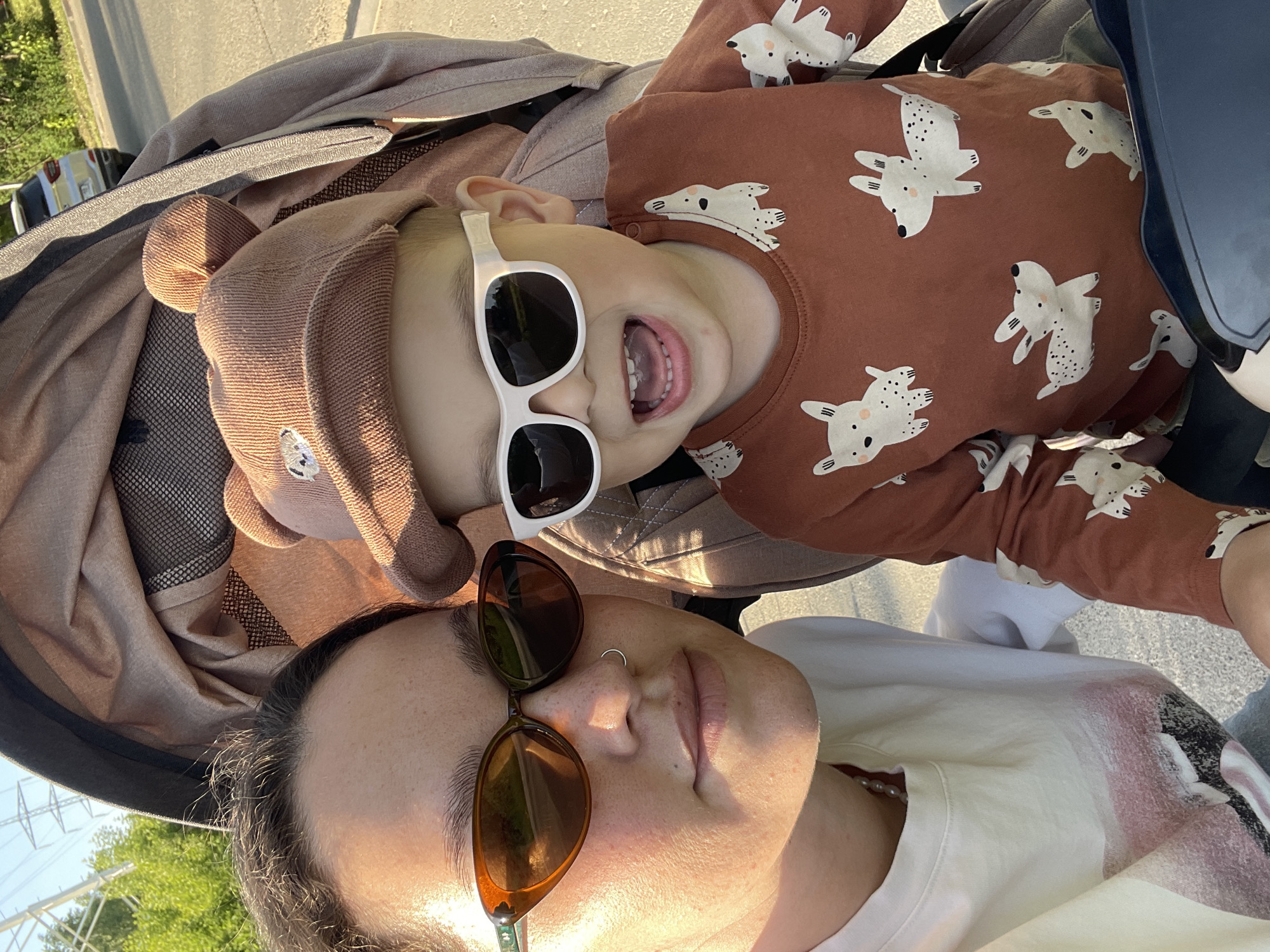 A selfie captured outdoors shows a woman and a toddler in a stroller. The woman, wearing brown sunglasses and a white t-shirt, is positioned at the bottom of the frame. The toddler, wearing white sunglasses and a brown bear-eared hat, is lying in a brown stroller, wearing a brown shirt with white animal prints. The sun is shining. Green foliage and power lines are visible in the background. The location is not identifiable as Izhevsk, Russia.