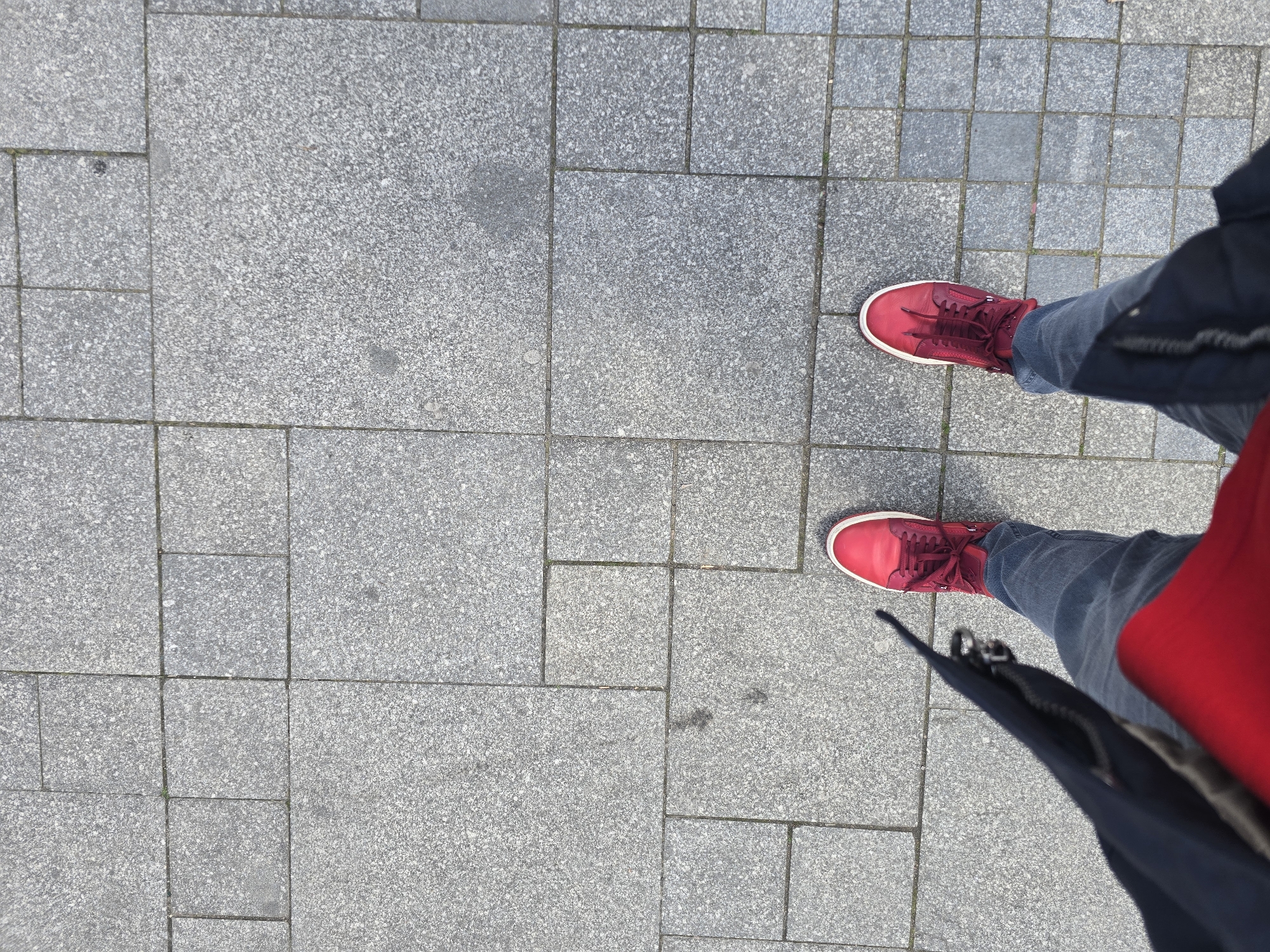 A high-angle shot shows a person standing on a paved surface. The paving consists of grey, rough-textured rectangular and square stones. The person wears jeans and bright red sneakers. Part of a dark blue or black jacket and a glimpse of a red shirt are visible, partially obscuring the view of the right leg and shoe. The setting appears to be a public walkway or square in a city, with a lack of other details to pinpoint the exact location within Sofia, Bulgaria. The focal point is on the person's footwear and the textured pavement.