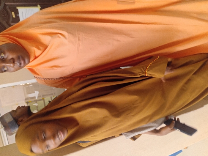 The image displays an indoor scene featuring three individuals. In the foreground, two females stand side-by-side. The female on the left, wearing a brown full-length abaya and matching hijab, holds a black smartphone in her right hand and has a light-colored strap over her left shoulder. Her face is slightly blurred, with her mouth partially open. The female on the right, wearing an orange full-length abaya and matching hijab, looks slightly upwards and to her left, with her mouth also partially open. A light-colored strap is visible on her left shoulder. In the blurred background, a male wearing a patterned cap and a light-colored shirt is visible in profile, looking to the right. The environment consists of light-colored walls, with a dark-framed doorway and a white sheet of paper or notice affixed to it visible in the background. The specific location within Kwazailewa, Nigeria, cannot be determined from the visual information presented.