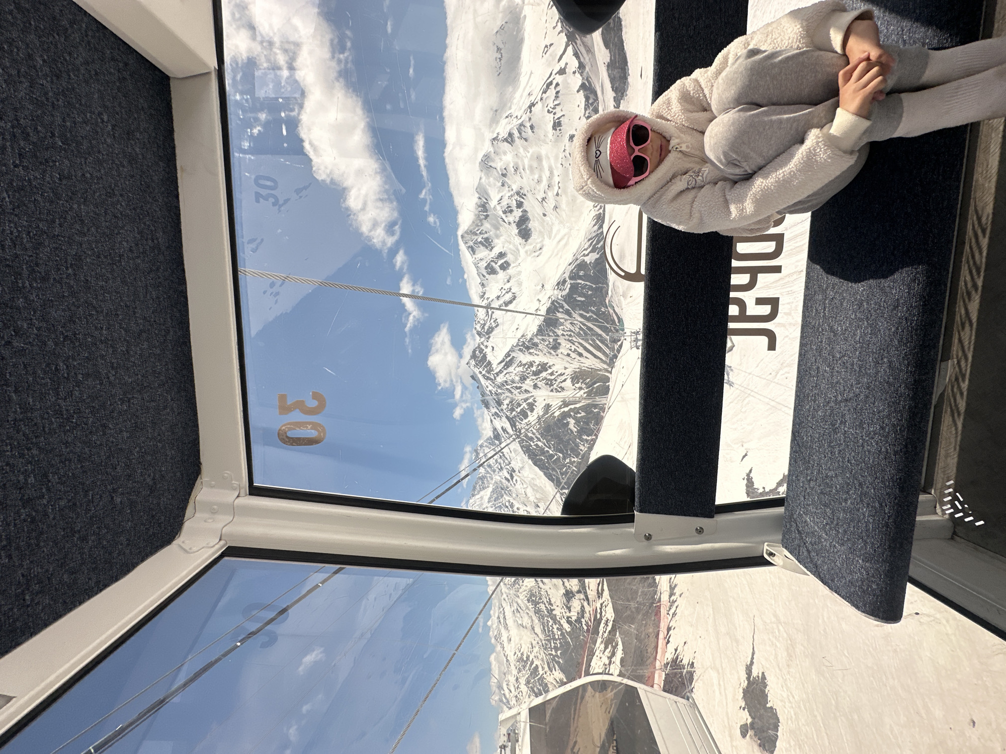 In Terskol, Russia, a young girl sits in a ski lift gondola, looking out at a snow-covered mountain range under a bright blue sky with scattered clouds. She is wearing a white hooded fleece jacket, gray sweatpants, and white socks. A pink baseball cap with a cat design and pink sunglasses adorn her head. The gondola interior features dark blue upholstered seats and a white frame. The window displays the number "30" in orange. Outside the window, ski lifts and a cable car are visible on the snowy slopes.
