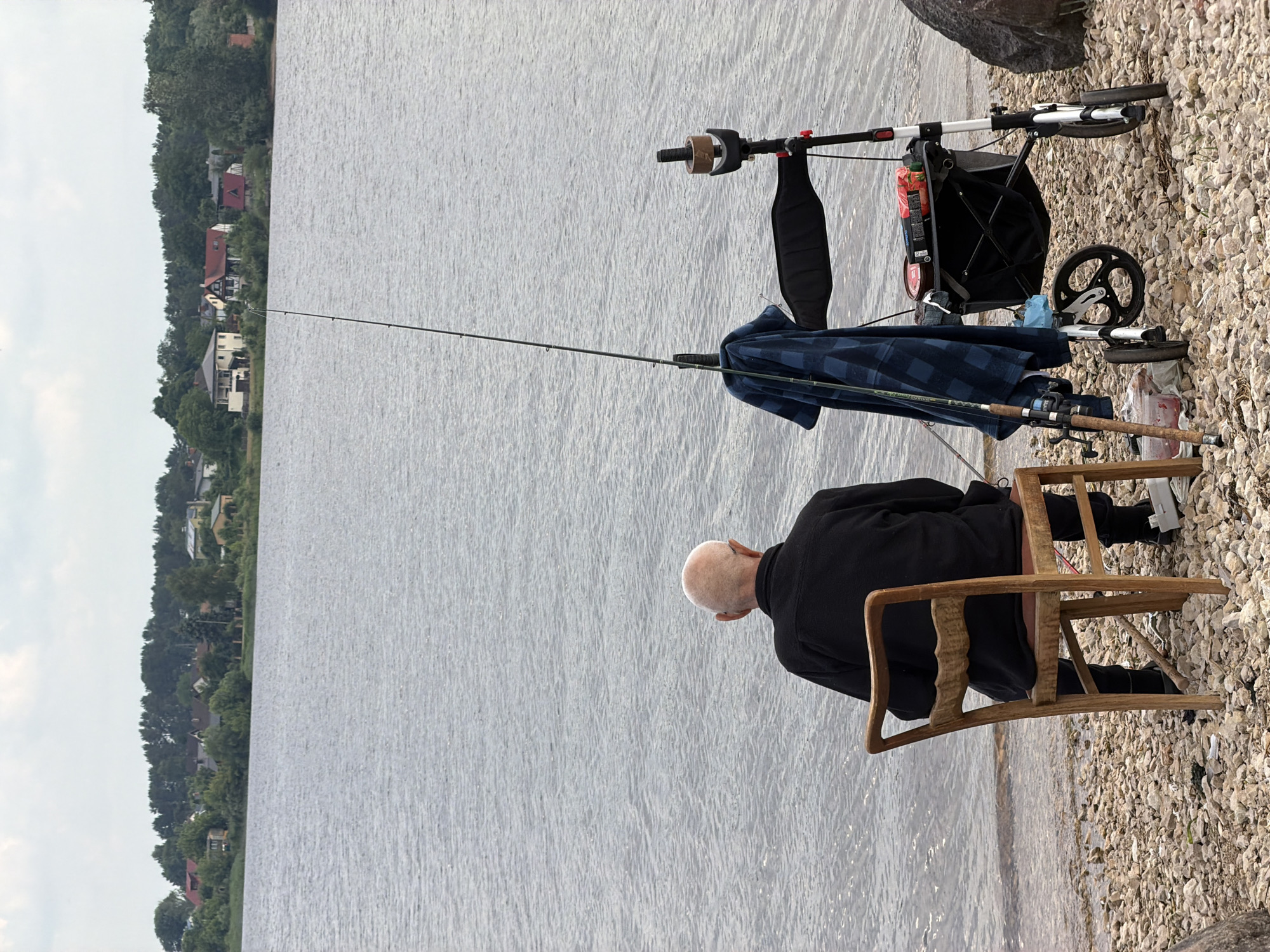 The image depicts an elderly bald man with glasses, wearing a black jacket and trousers, sitting on a wooden chair by the shore of a large body of water. He is facing away from the camera, looking out at the water. To his right is a walker with a basket, which holds fishing supplies including tackle boxes and bait. Two fishing rods are propped up next to the walker and the chair. The ground is covered in small stones. In the background, across the water, is a shoreline with trees and houses. The sky is overcast. The scene appears to be a quiet, outdoor fishing spot. The location is not specified in the image.