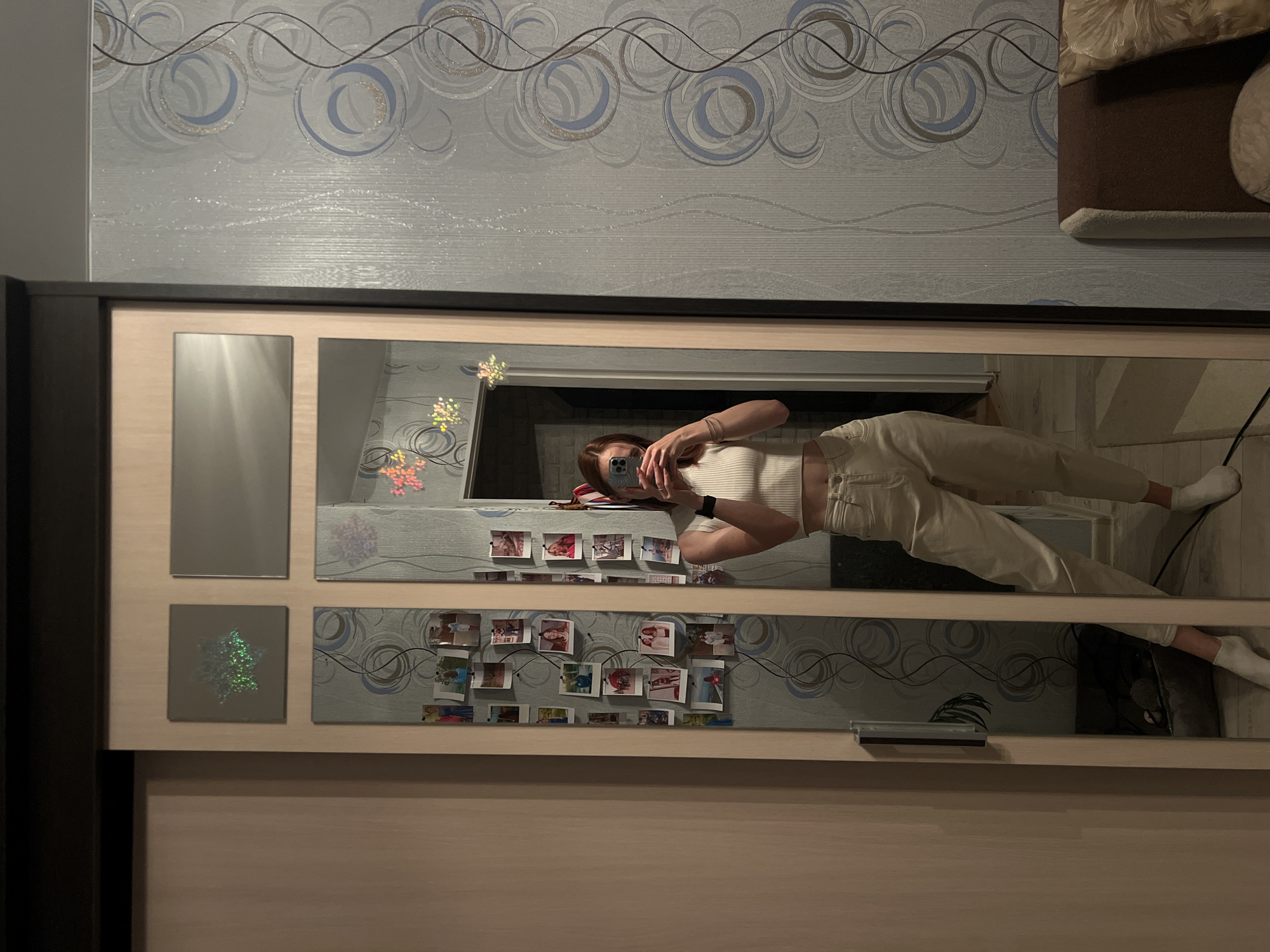 A young woman stands in front of a full-length mirror in what appears to be a bedroom in Chaikovsky, Russia. She is wearing a white crop top and light-colored pants. The mirror is part of a large wardrobe with sliding doors. The wall behind the mirror is decorated with a pattern of blue and grey circles. Several framed photographs are attached to the wardrobe doors, near the mirror. The room is lit by artificial light. The woman is holding a phone and taking a selfie. The overall impression is one of a casual home environment.