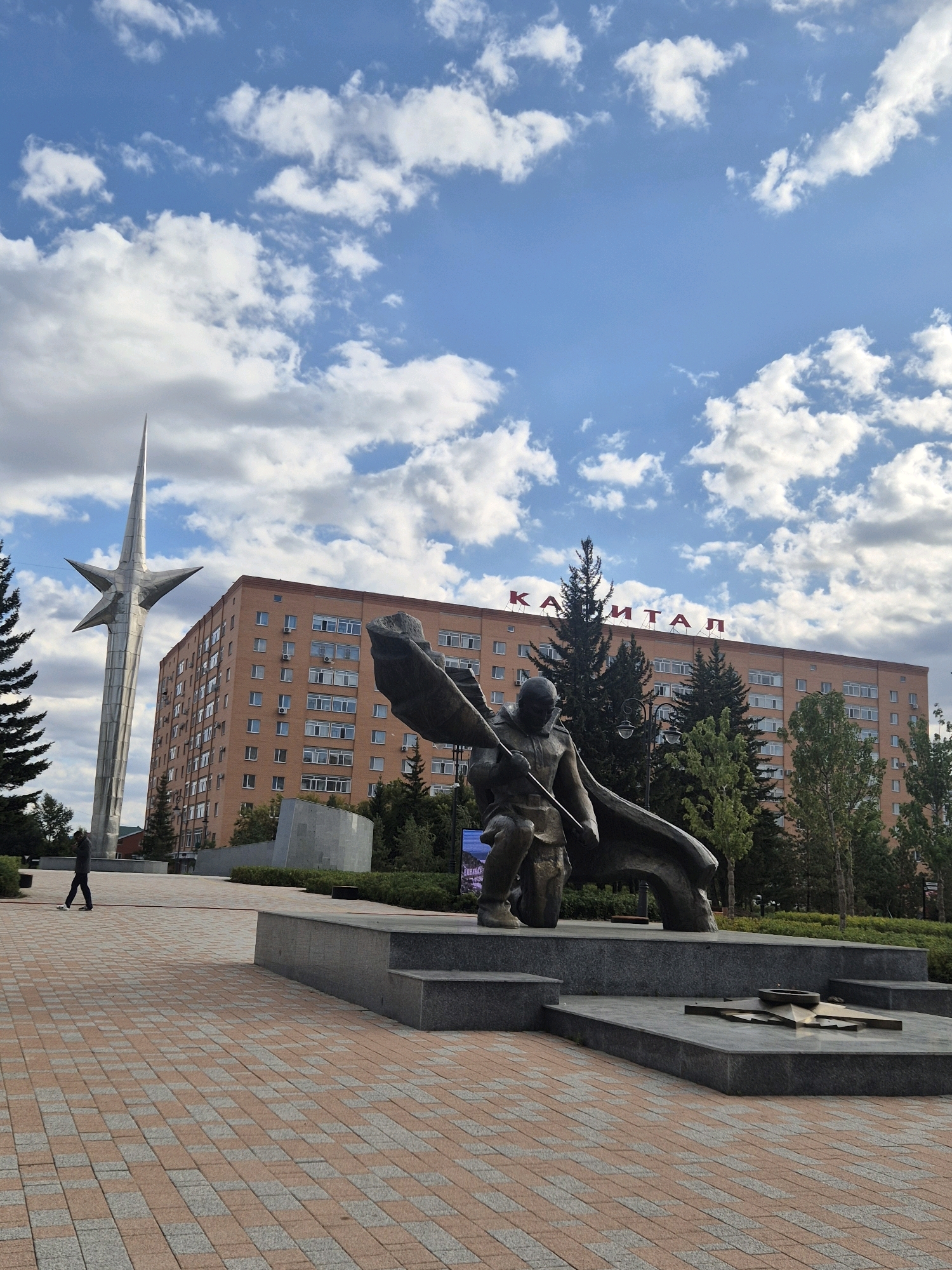 The image shows a monument in Kokshetau, Kazakhstan, with a tall, stylized star sculpture on the left and a bronze statue of a figure holding a flag in the foreground. Behind the monument, a multi-story brick building with "КАПИТАЛ" (Kapital) written in red letters on its facade dominates the background. The scene is set on a paved plaza with a pattern of orange and gray bricks. A single person is walking on the plaza in the distance. The sky is blue with scattered white clouds. The environment appears to be an urban public space.