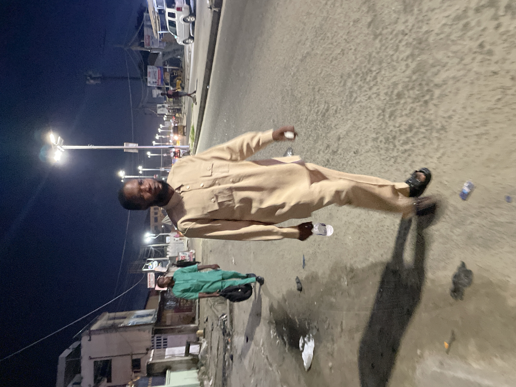The image depicts an outdoor scene in Ado, Nigeria, at night. The scene is lit by streetlights and some ambient lighting from nearby businesses. The pavement is gray and appears to be relatively clean in some areas, but littered with debris in others.
Two men are visible in the foreground. The first man is dressed in a light brown, long-sleeved traditional attire. He is holding an object that appears to be a small bottle or a container in his hand. He is wearing black sandals. The second man is wearing a green shirt and pants and has a black bag slung over his shoulder.
In the background, a line of buildings and businesses is visible, with various signs and advertisements displayed. A white SUV is parked on the side of the road, and another person is walking along the sidewalk. Electrical wires stretch across the top of the frame.