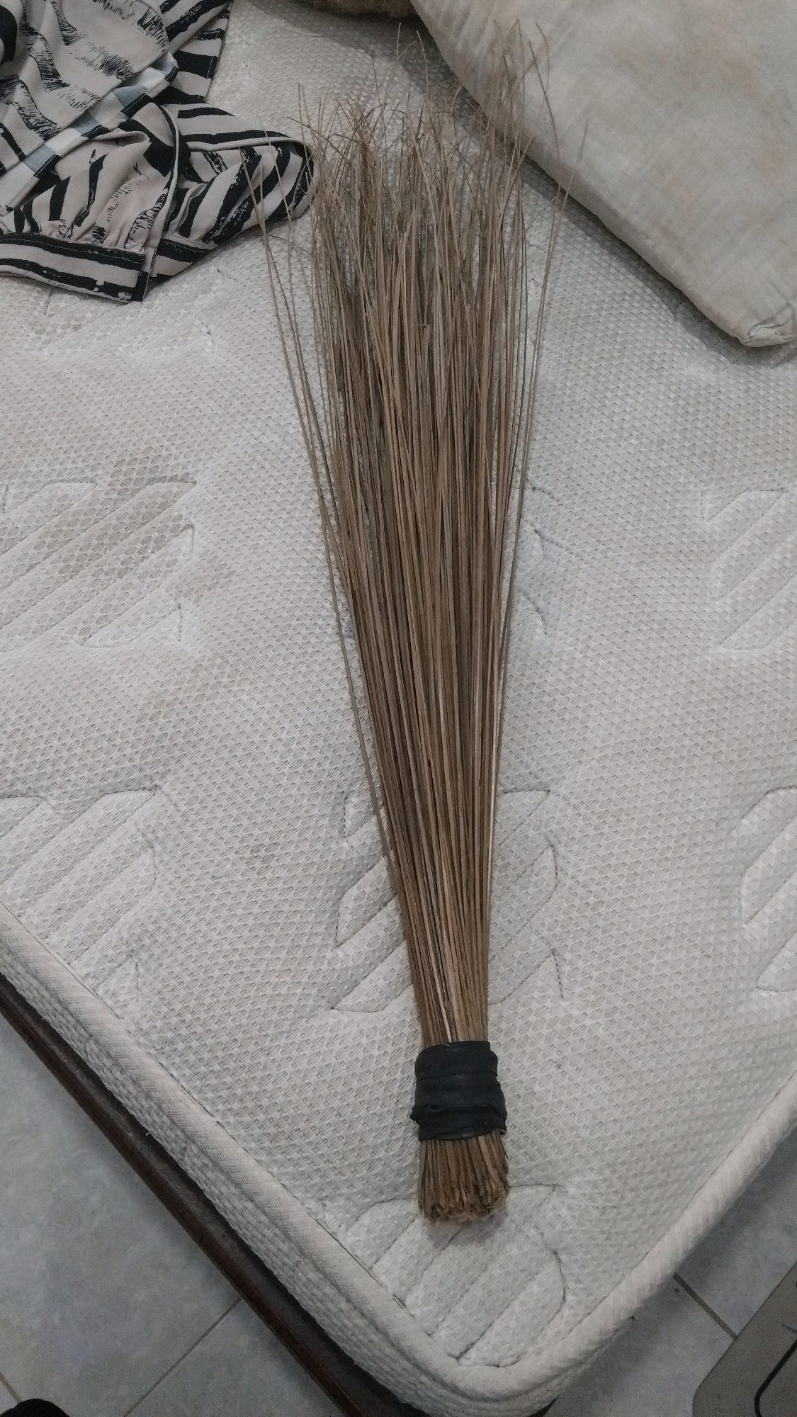 A handmade broom made of dried coconut fronds is lying on a white textured mattress. The broom is bound with a black elastic band. To the left of the broom, a folded piece of fabric with a black and white striped pattern is visible. In the upper right corner, a light-colored pillow is partially in view. The scene appears to be indoors, possibly a bedroom, in the city of Kebon IX, Indonesia.
