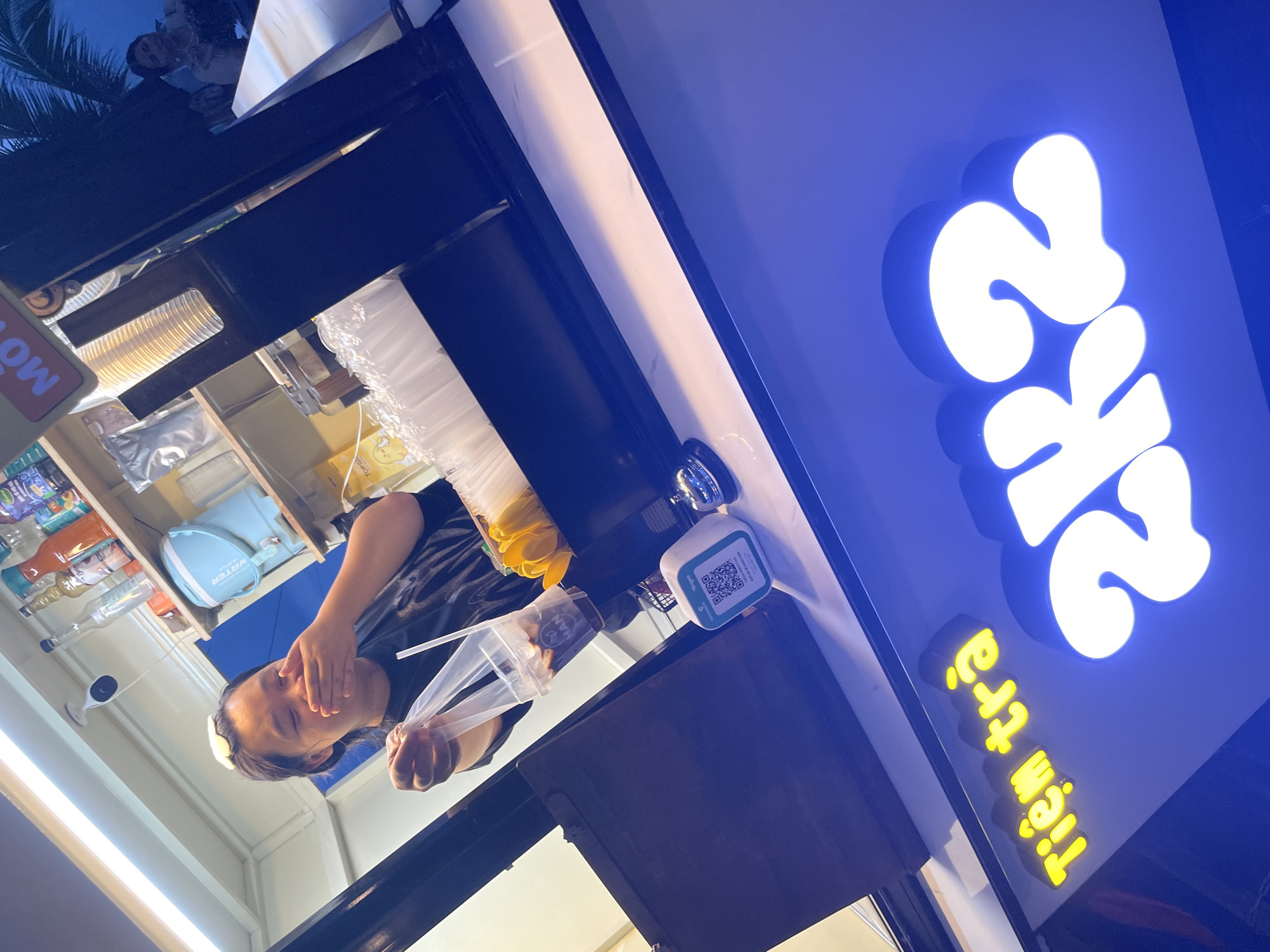 In Mũi Né, Vietnam, a person is standing behind a counter in what appears to be a beverage stall or shop. The person is wearing a dark t-shirt and has their hand over their mouth, seemingly in a gesture of surprise or amusement. They are holding a clear plastic cup filled with a dark liquid and ice, with a straw inserted. The cup is partially enclosed in a clear plastic bag.
Behind the person, shelves are visible, stocked with various bottles of syrups and other beverage ingredients, as well as a light blue appliance. A CCTV camera is mounted on the ceiling. On the counter, a white and blue QR code payment terminal is present, along with a small silver bell.
In the foreground, a brightly lit blue sign with yellow lettering that reads "Tiệm trà" is prominently displayed, followed by large white illuminated numbers and letters that appear to spell "2K2". The overall environment suggests a commercial establishment, possibly a bubble tea shop or a similar refreshment vendor. A palm tree is visible in the background, hinting at a coastal or tropical location.