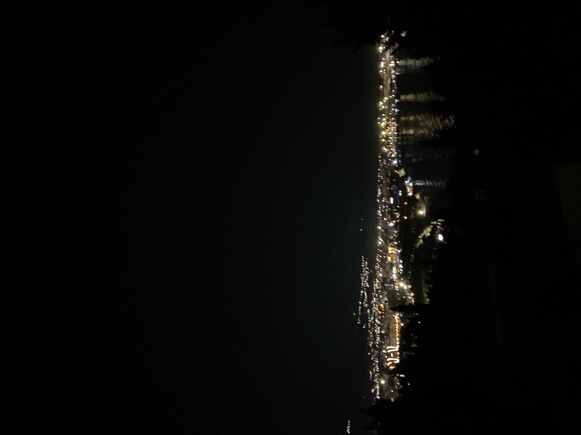 The image is an eye-level, nighttime view of the city of Šušanj, Montenegro, facing east. The city lights are visible along the coastline, reflected in the dark water. There are no people visible in the image.