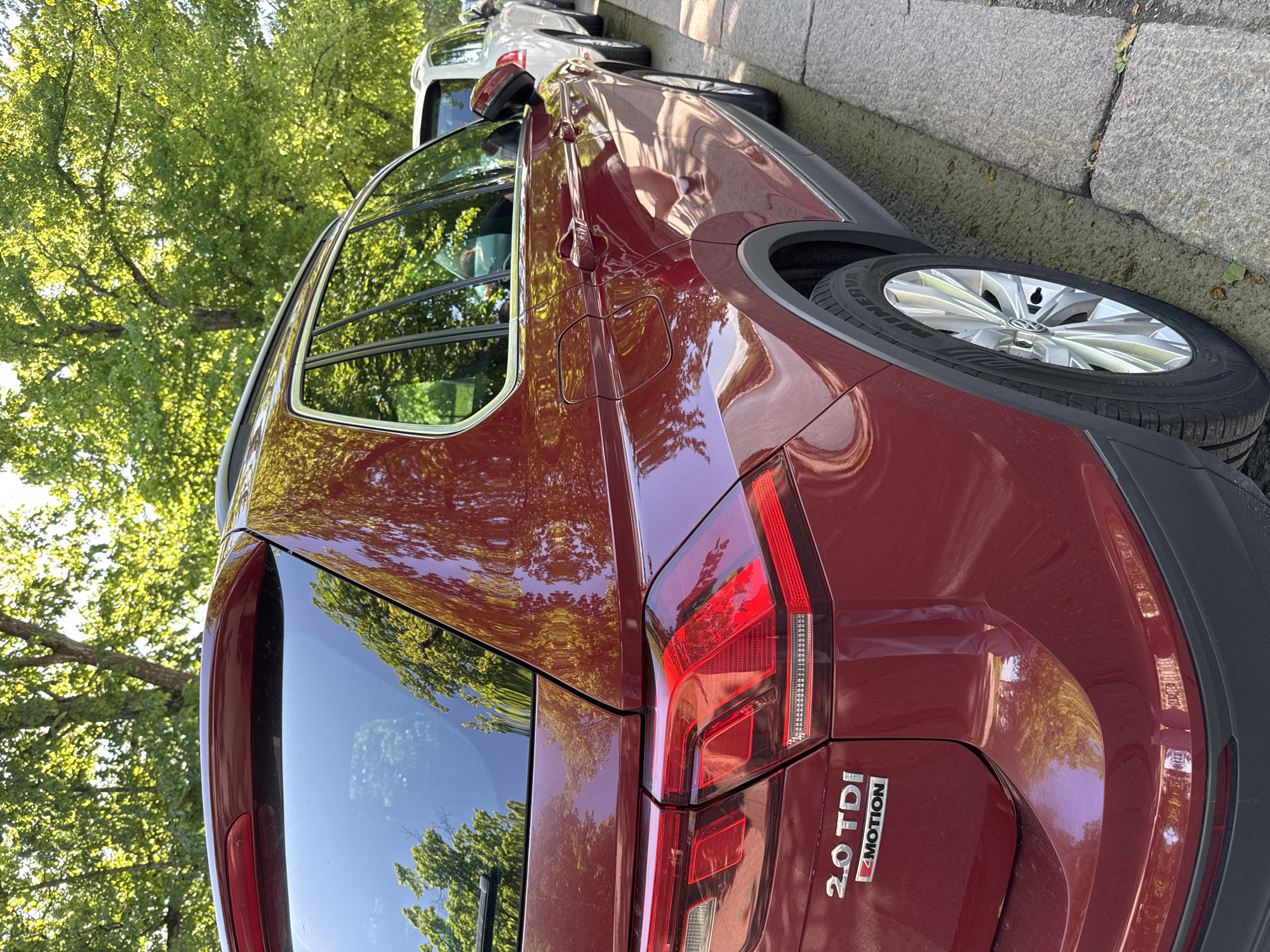 A dark red Volkswagen Tiguan SUV (identified by "2.0 TDI" and "4MOTION" badging) is parked parallel to a stone curb on the right side of a street. The vehicle's rear and right side are visible. It features silver multi-spoke alloy wheels with Minerva brand tires, LED taillights, a rear wiper, and roof rails. The car's glossy finish reflects numerous bright green tree leaves, indicating a sunny environment. A faint reflection of a person wearing a light-colored face mask is visible in the driver's side rear window. A white SUV is parked directly behind the red Volkswagen. The setting is an urban or suburban street lined with mature trees. The location is in the city of Unknown, Unknown.