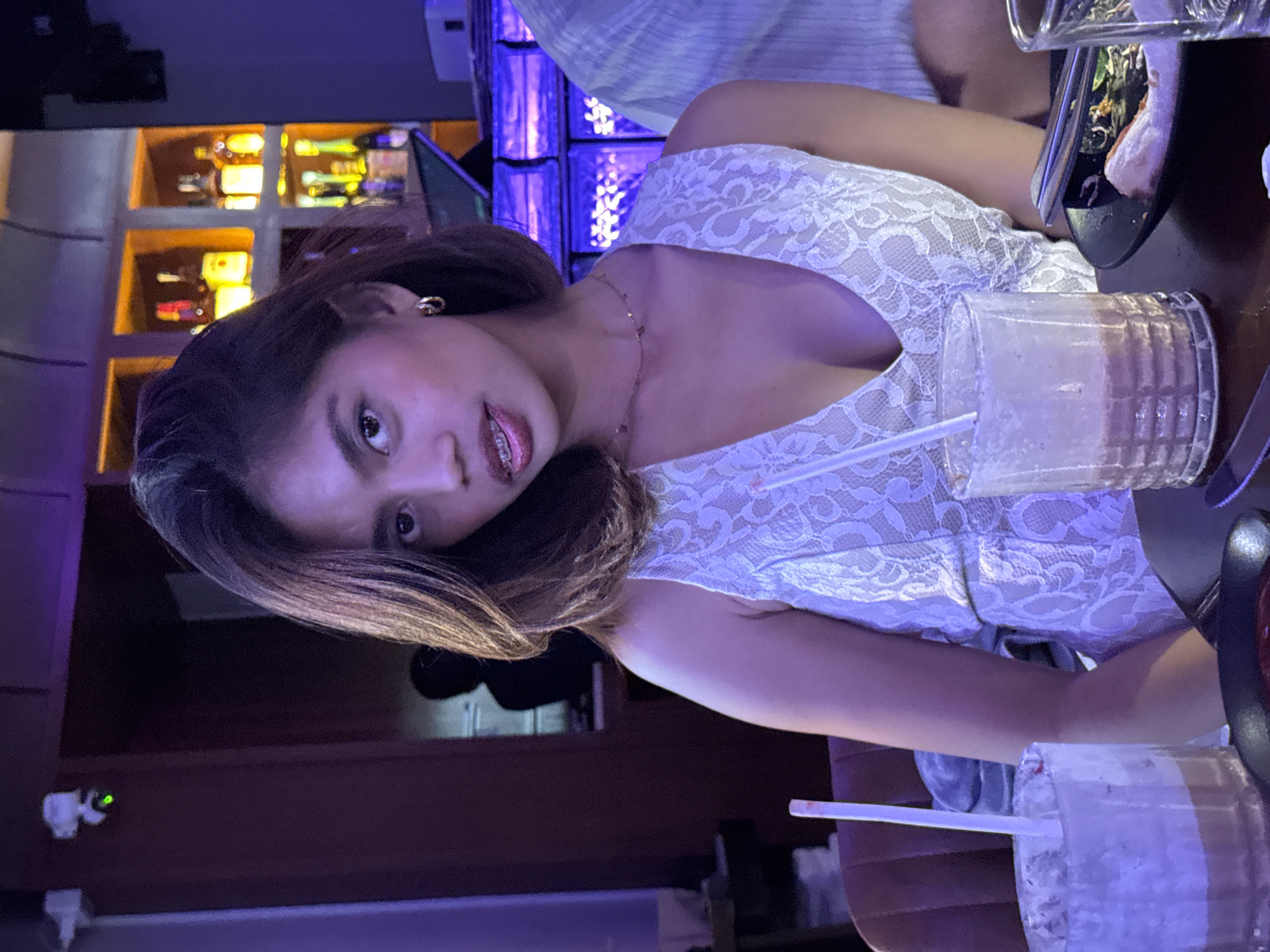 A young woman with dark hair, wearing a white lace dress and braces, is seated at a table in a dimly lit establishment. She is looking towards the camera with a neutral expression. In front of her is a glass with a beverage and a straw. The background shows shelves with bottles and purple lighting. The scene appears to be in a bar or restaurant.