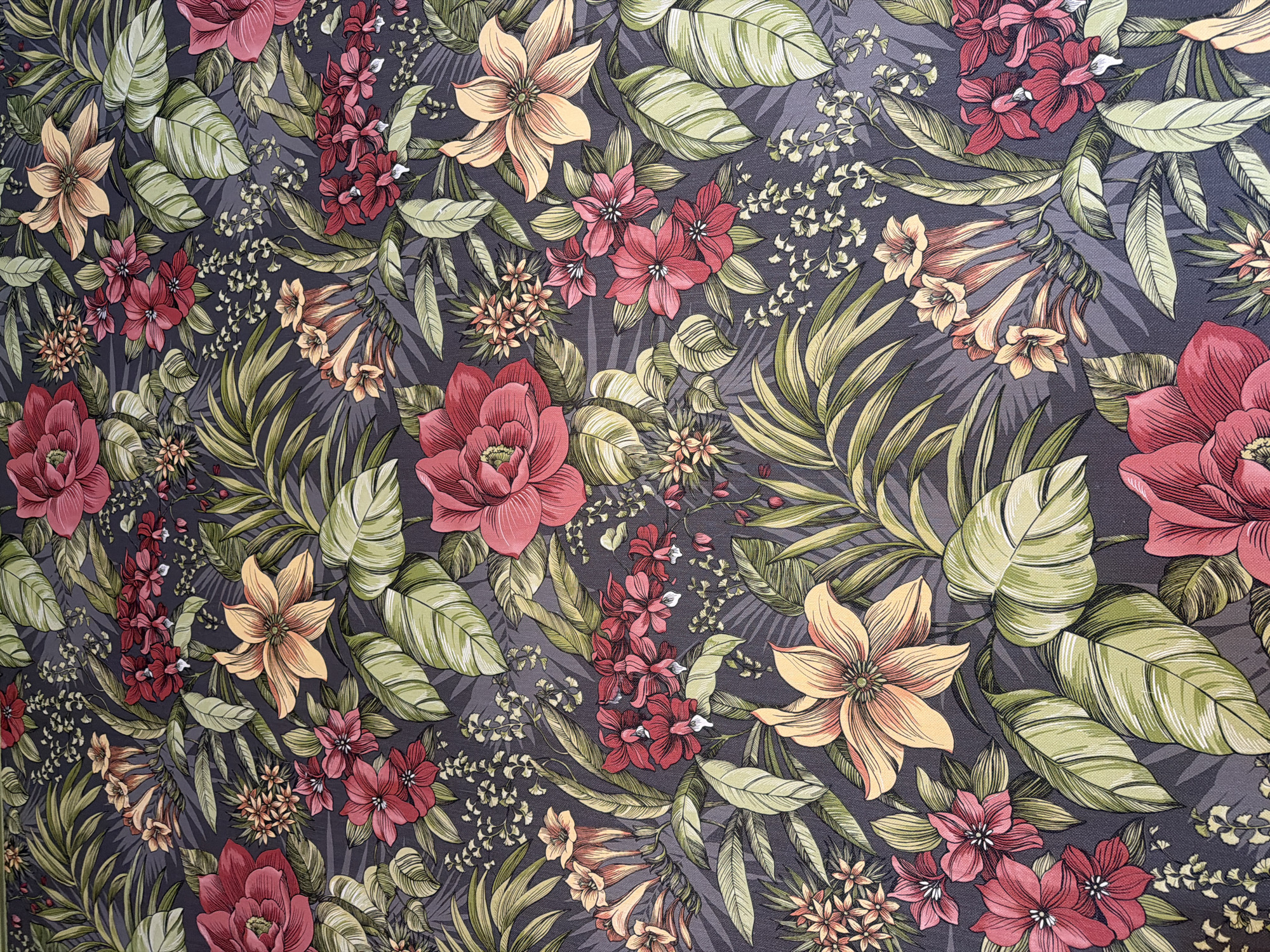 This is a top-down, eye-level, full shot of a floral fabric pattern with a dark gray background. The pattern features a variety of tropical flowers and leaves rendered in shades of red, pink, yellow, and green, with some accents of orange. Prominent elements include large, open red and pink flowers with detailed petals, smaller clusters of pink and red flowers, and trumpet-shaped yellow and orange flowers. Various large, broad green leaves with lighter green veins are interspersed throughout the composition, along with spiky, elongated green leaves that resemble palm fronds. Smaller, delicate clusters of light green and yellow foliage are also visible. The overall impression is a dense and vibrant botanical design.
